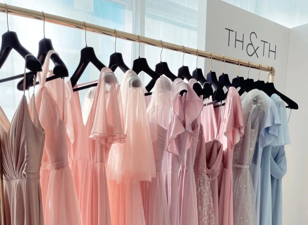 A clothing rack displays an array of elegant pastel pink and blush dresses on black hangers in front of a window, with a “TH&TH” sign in the background.