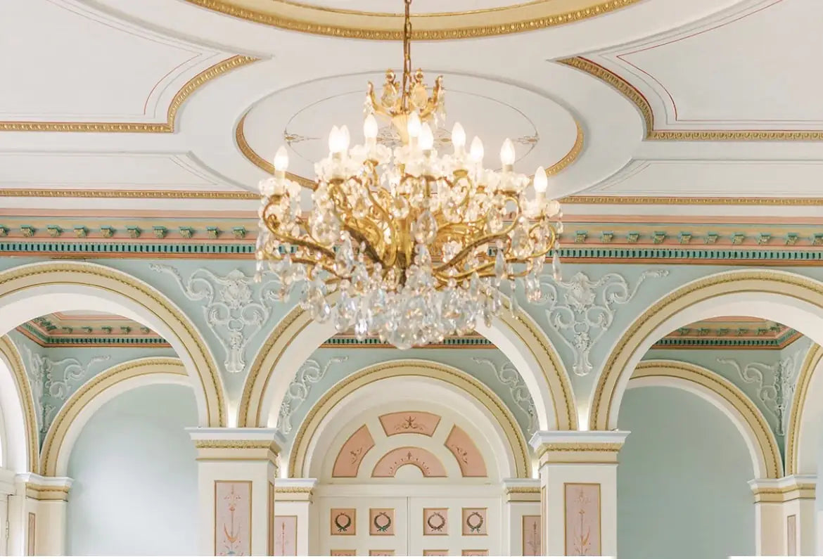 An ornate crystal chandelier hangs from a decorated ceiling with gold accents, intricate moldings, and pastel green and cream arched walls in an elegant, classical-style room.