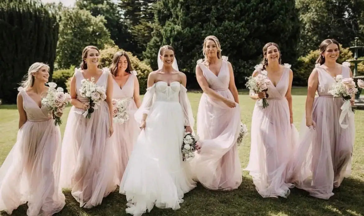 A bride in a white dress walks outside on grass with six bridesmaids in light pink dresses, all holding bouquets and smiling, surrounded by greenery and trees.