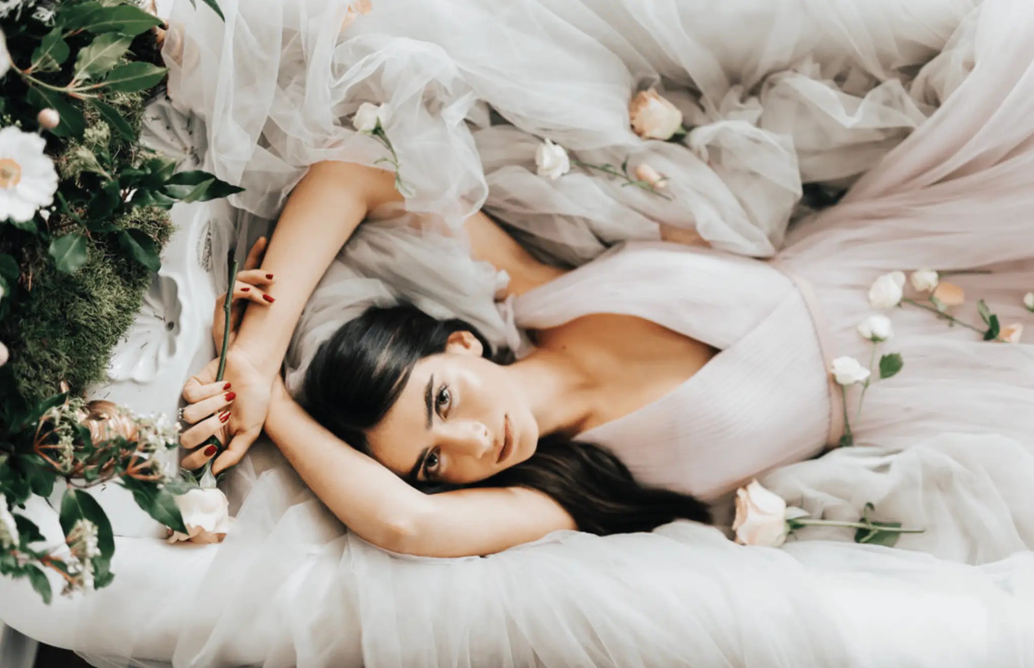A woman in a flowing, pale pink dress lies on soft, sheer fabric, surrounded by white and blush roses and greenery. She gazes upward with long brown hair spread around her, creating a dreamy, elegant scene.
