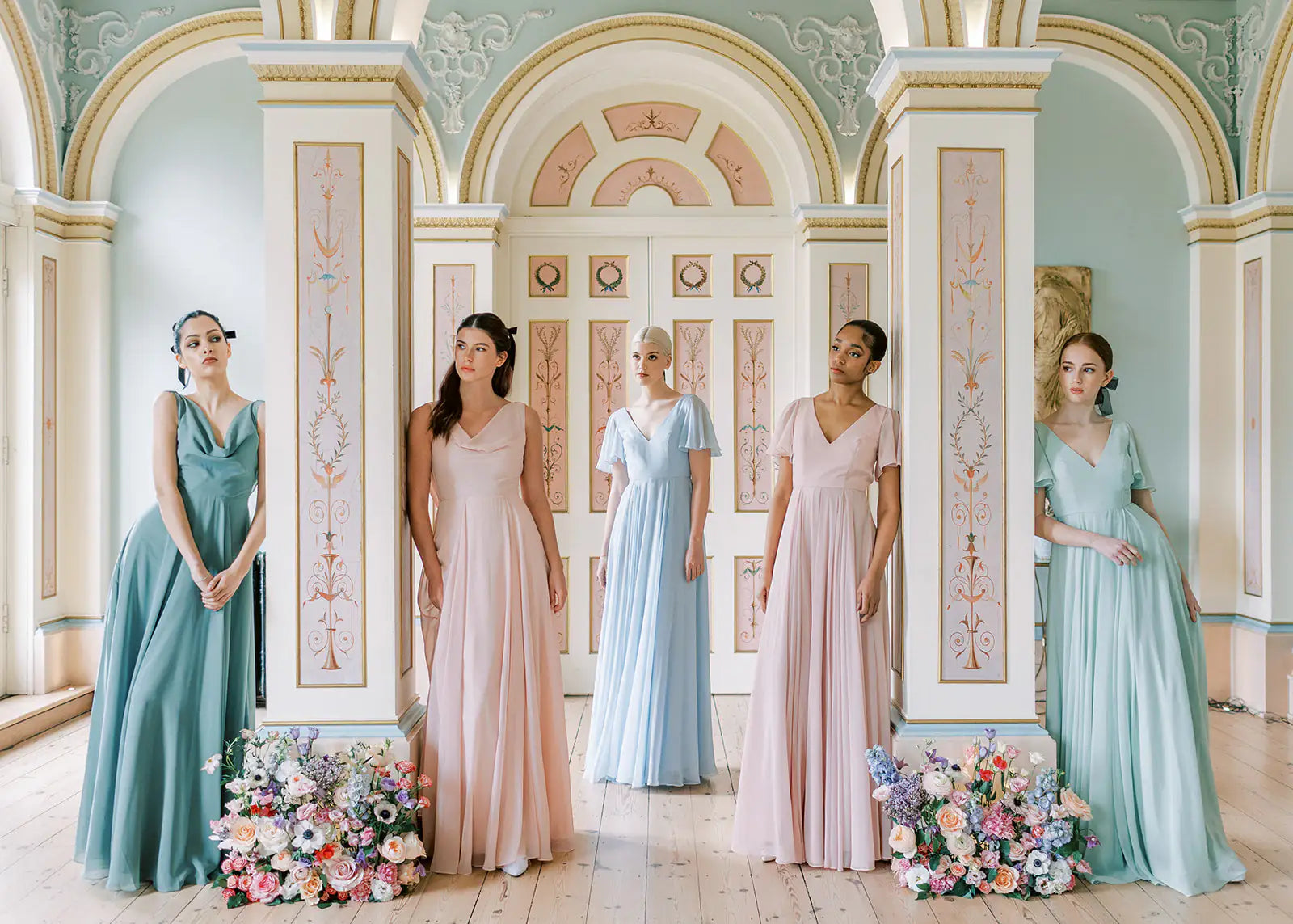 Five women in pastel-colored gowns pose elegantly in a bright, ornate room with high arches. Floral arrangements sit on the floor beside them, complementing the classical decor and soft, airy atmosphere.
