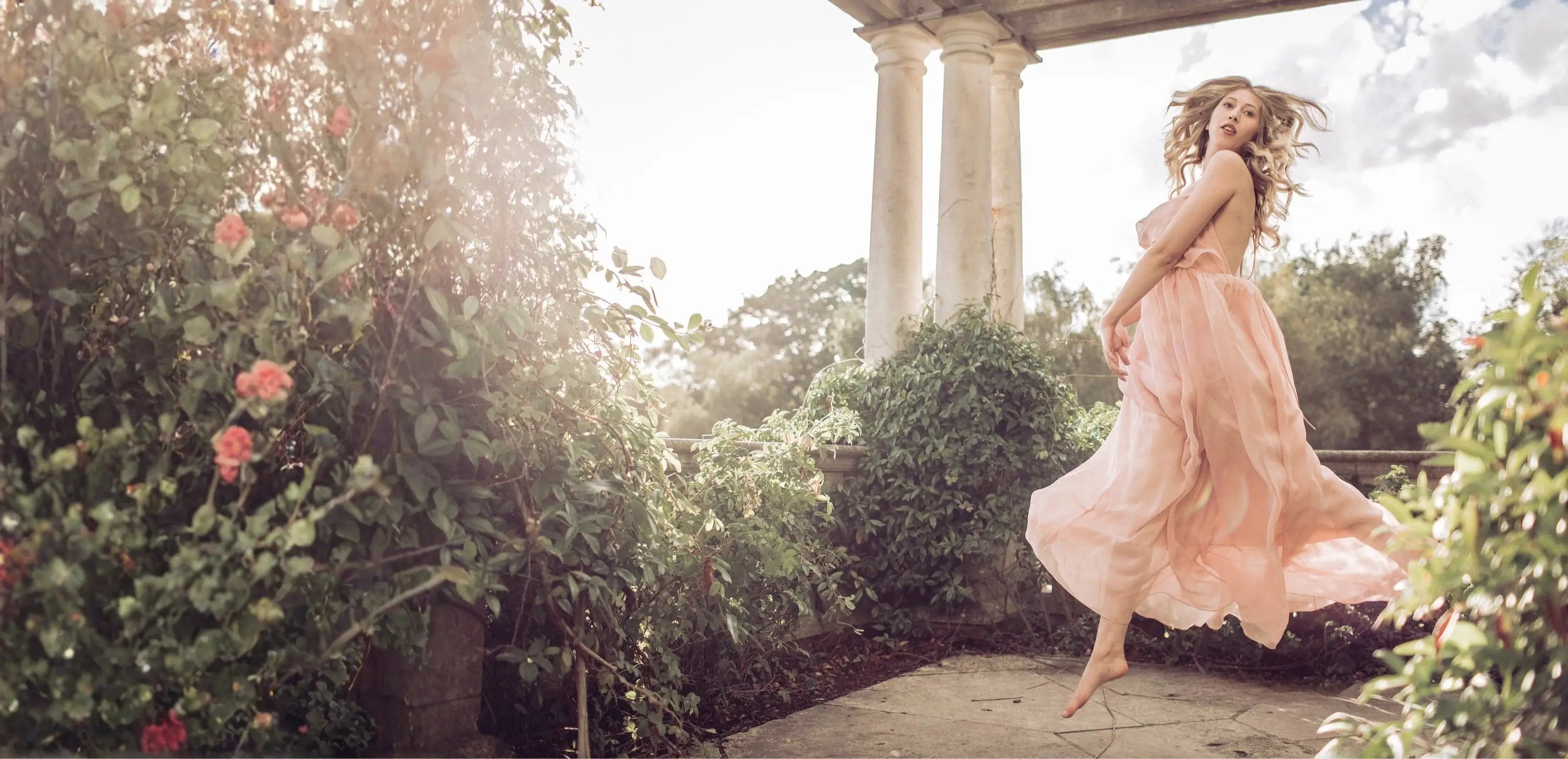 A woman in a flowing pink dress jumps gracefully near blooming rose bushes by stone columns under a partly cloudy sky, with sunlight streaming through the garden.