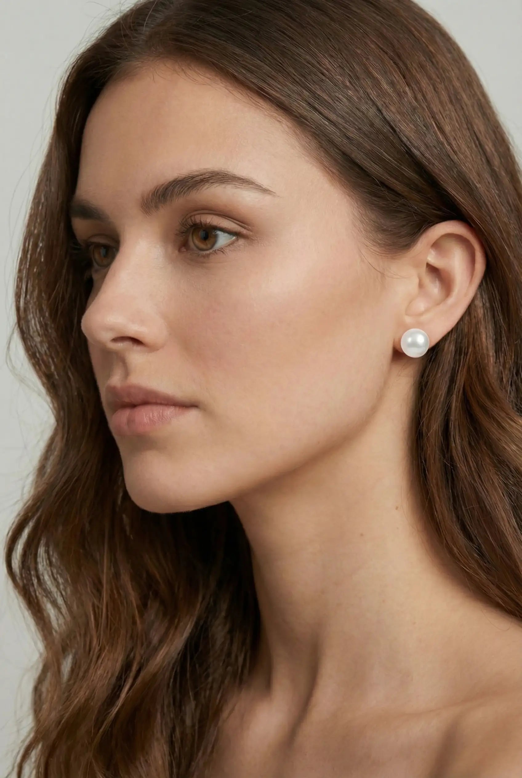 A woman with long brown hair and natural makeup wears TH&TH 10mm Pearl Stud Earrings, highlighting the classic elegance of these bridal earrings against a plain, light background.