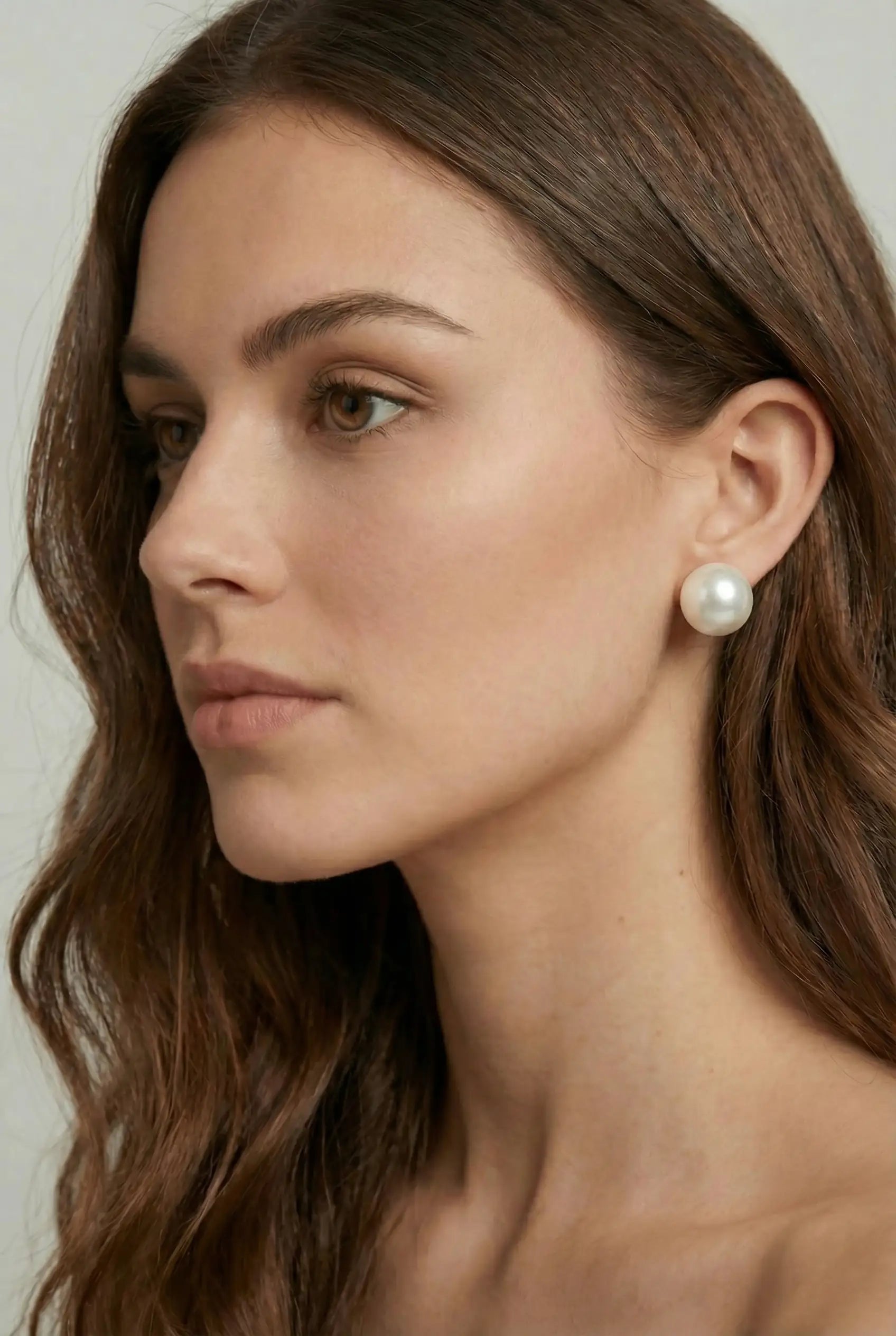 A woman with long brown hair, shown in profile and wearing TH&TH's 12mm Pearl Stud Earrings, displays an elegant bridal style. Her clear, smooth skin and neutral expression highlight the classic appeal of these statement earrings.