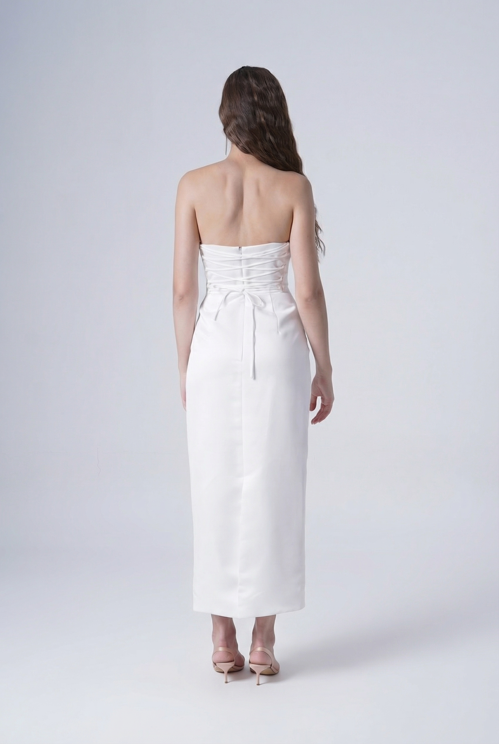 A woman with long wavy hair faces away, wearing the TH&TH Ivy Bridal Dress in Ivory and nude heels, set against a plain light background.
