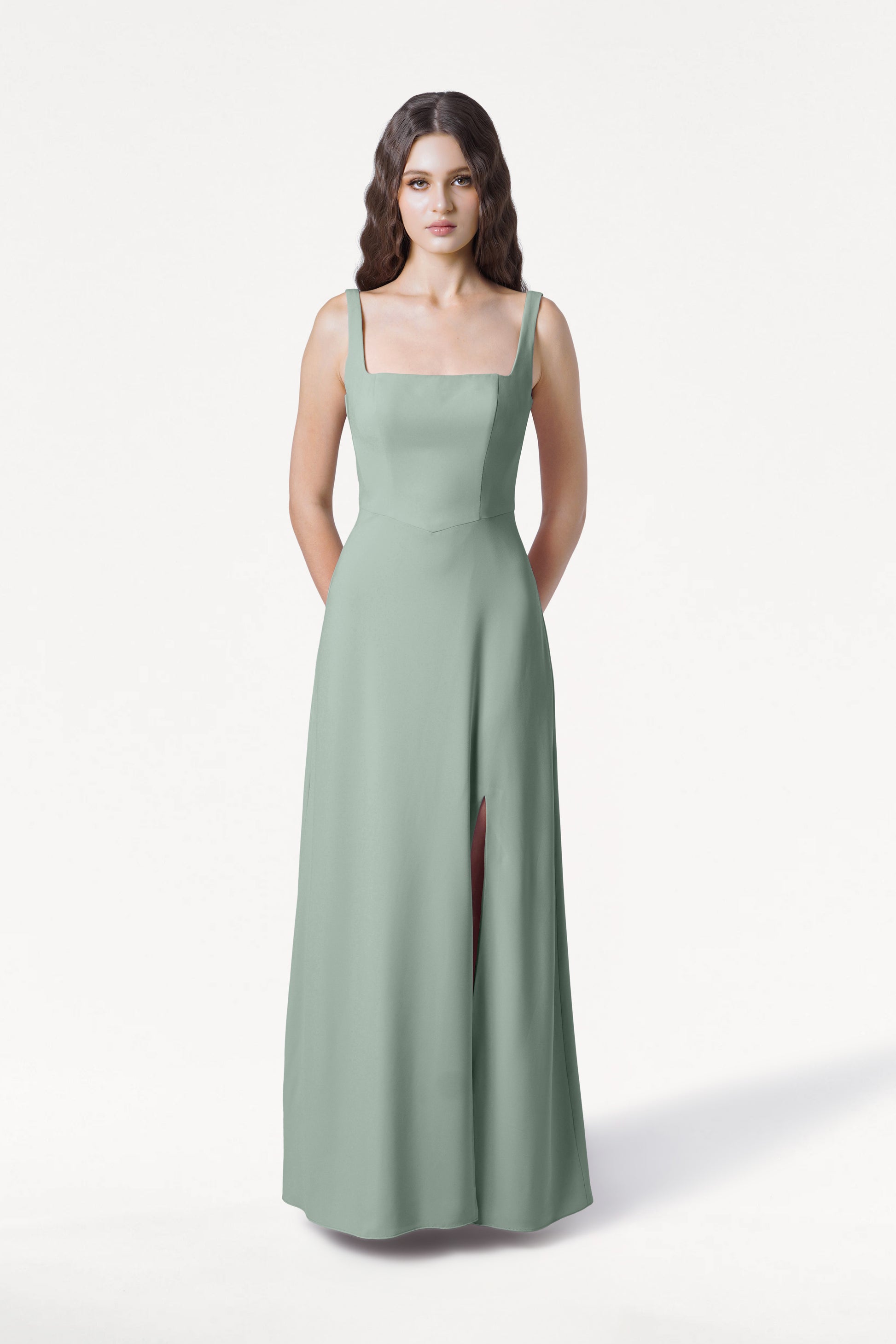 A woman with long, wavy dark hair wears the TH&TH Helena Bridesmaid Dress in Sage Green, featuring a sleeveless moss crepe design with a square neckline and high slit, standing against a plain white background.