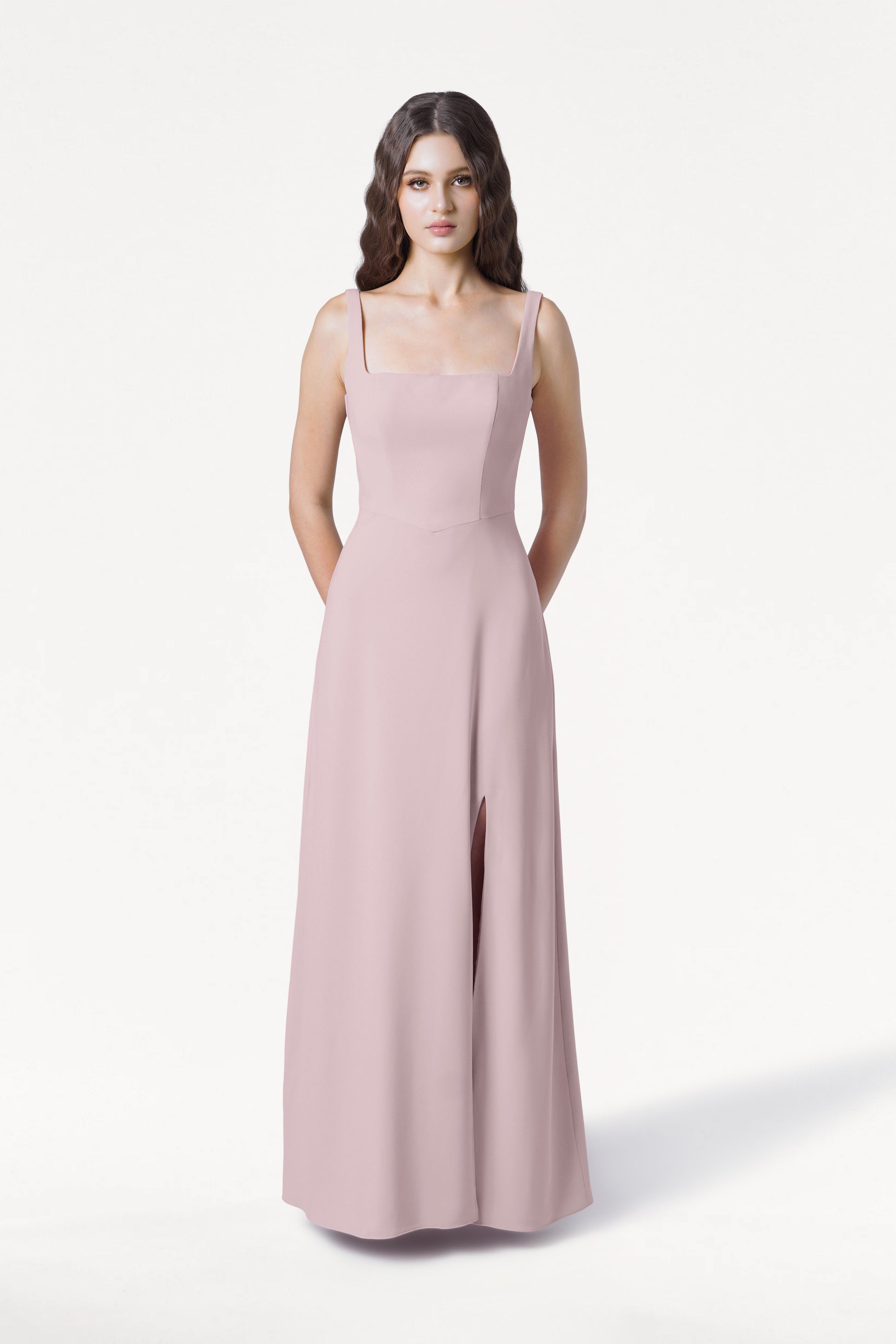 A woman with long brown hair wears the TH&TH Helena Bridesmaid Dress in Smoked Blush—a sleeveless, floor-length gown with a square neckline and front slit—standing facing forward against a plain white background.