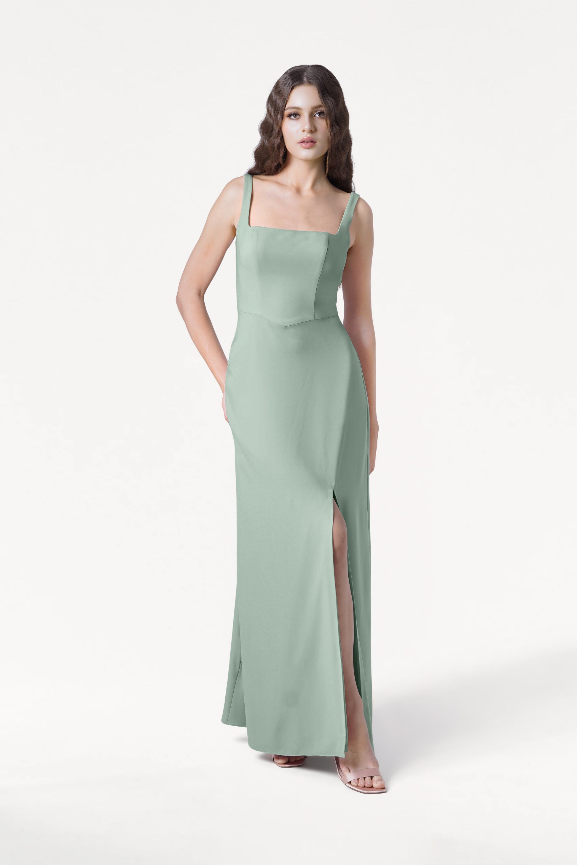 A woman in the TH&TH Helena Bridesmaid Dress in sage green—sleeveless, floor-length moss crepe with a square neckline and high slit—stands against a plain white background.