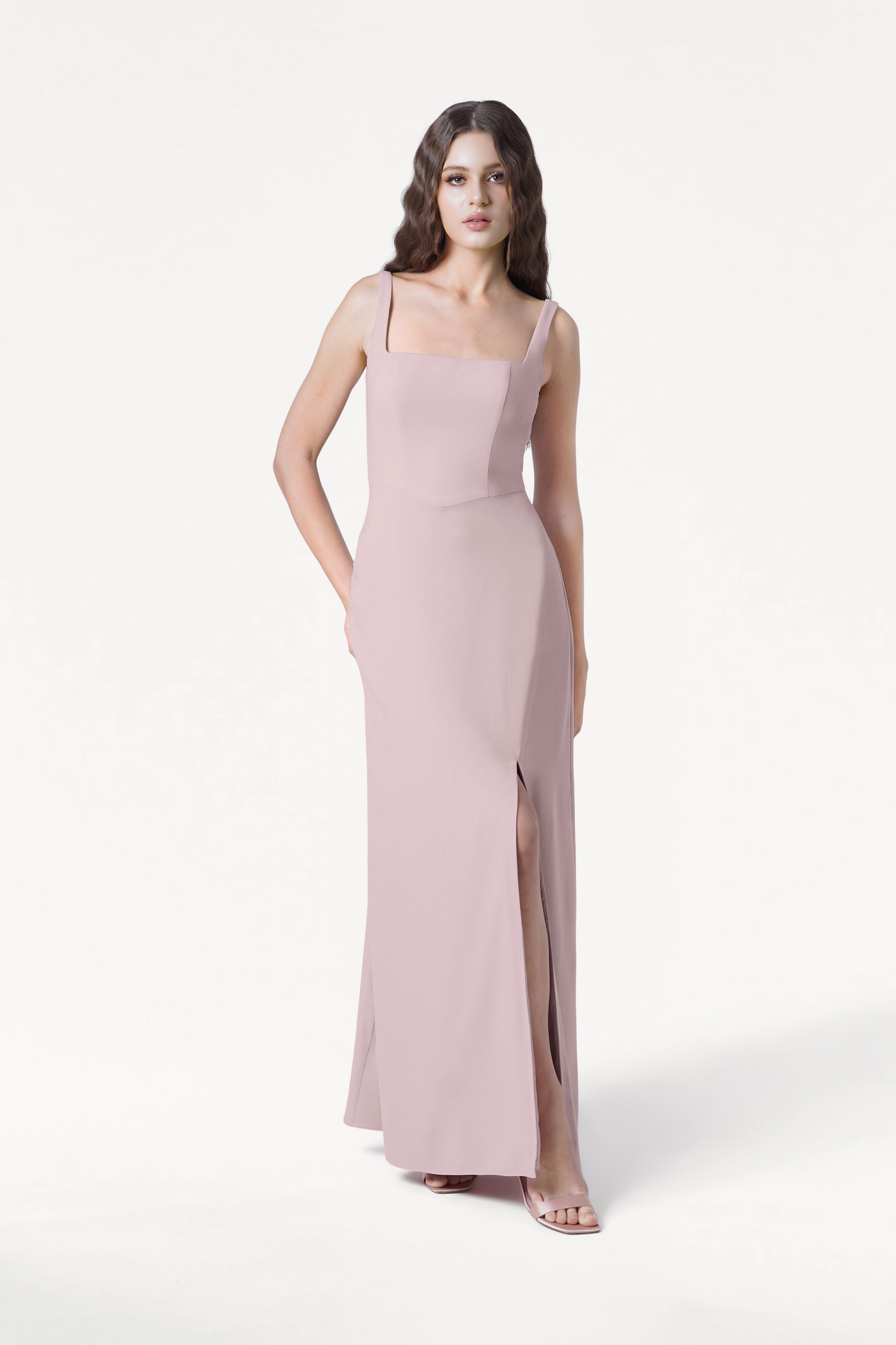 A woman poses confidently with one hand in her pocket, wearing the TH&TH Helena Bridesmaid Dress in Smoked Blush—a modern moss crepe gown featuring a square neckline and thigh-high slit—set against a plain white background.