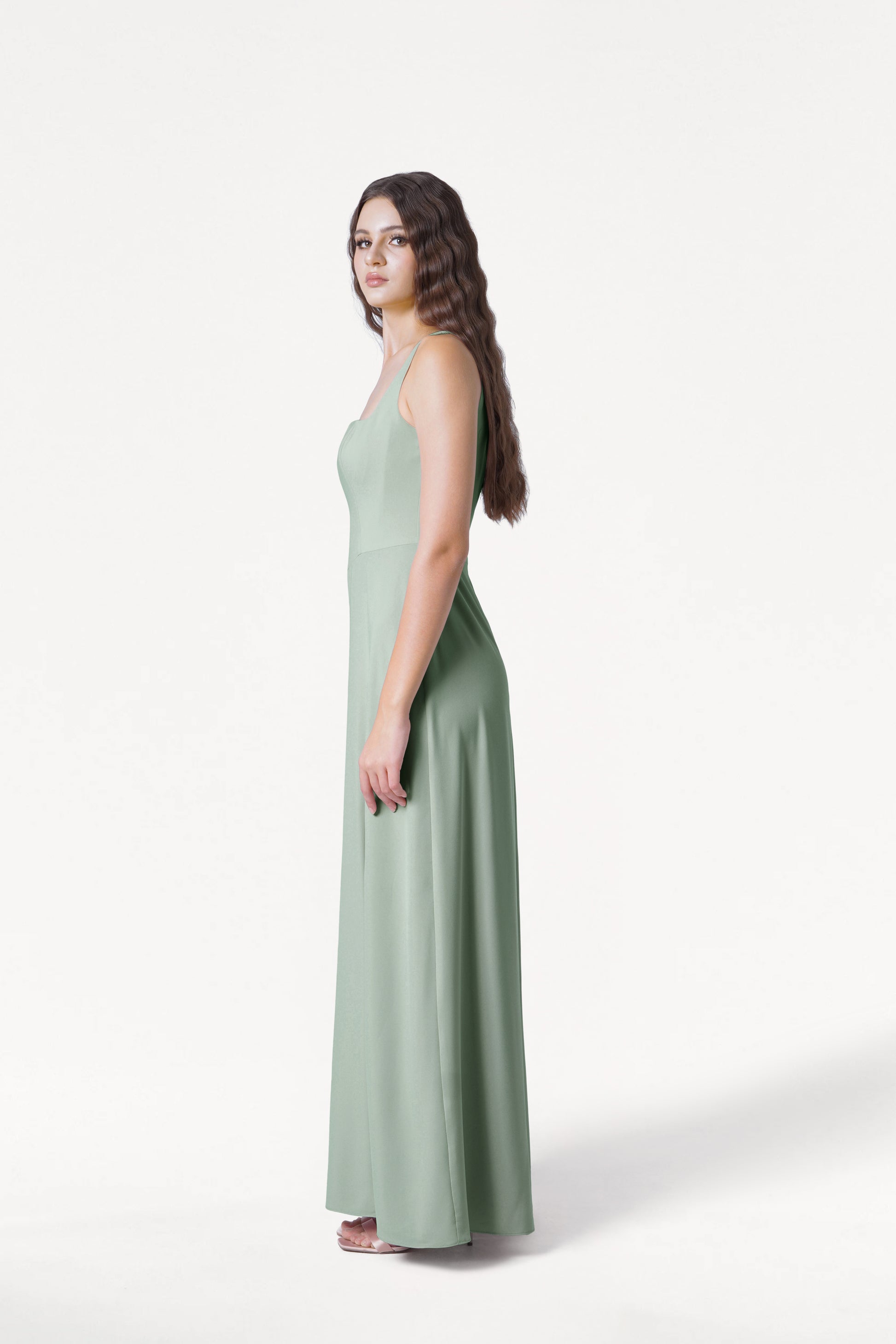 A woman with long wavy hair stands in profile wearing the TH&TH Helena Bridesmaid Dress in Sage Green with white sandals, posed against a plain white background.
