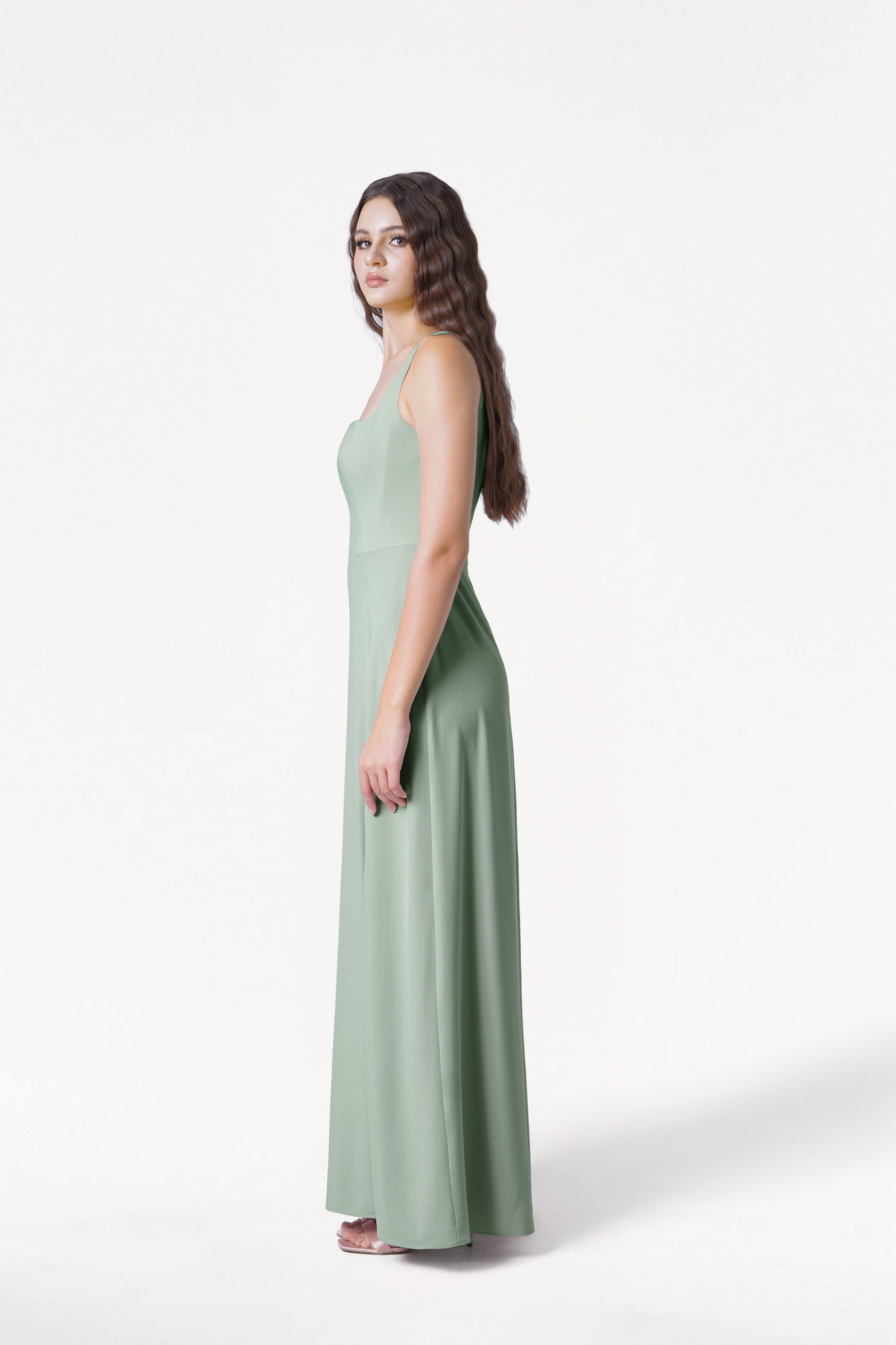A woman with long wavy hair stands in profile wearing the TH&TH Helena Bridesmaid Dress in Sage Green with white sandals, posed against a plain white background.
