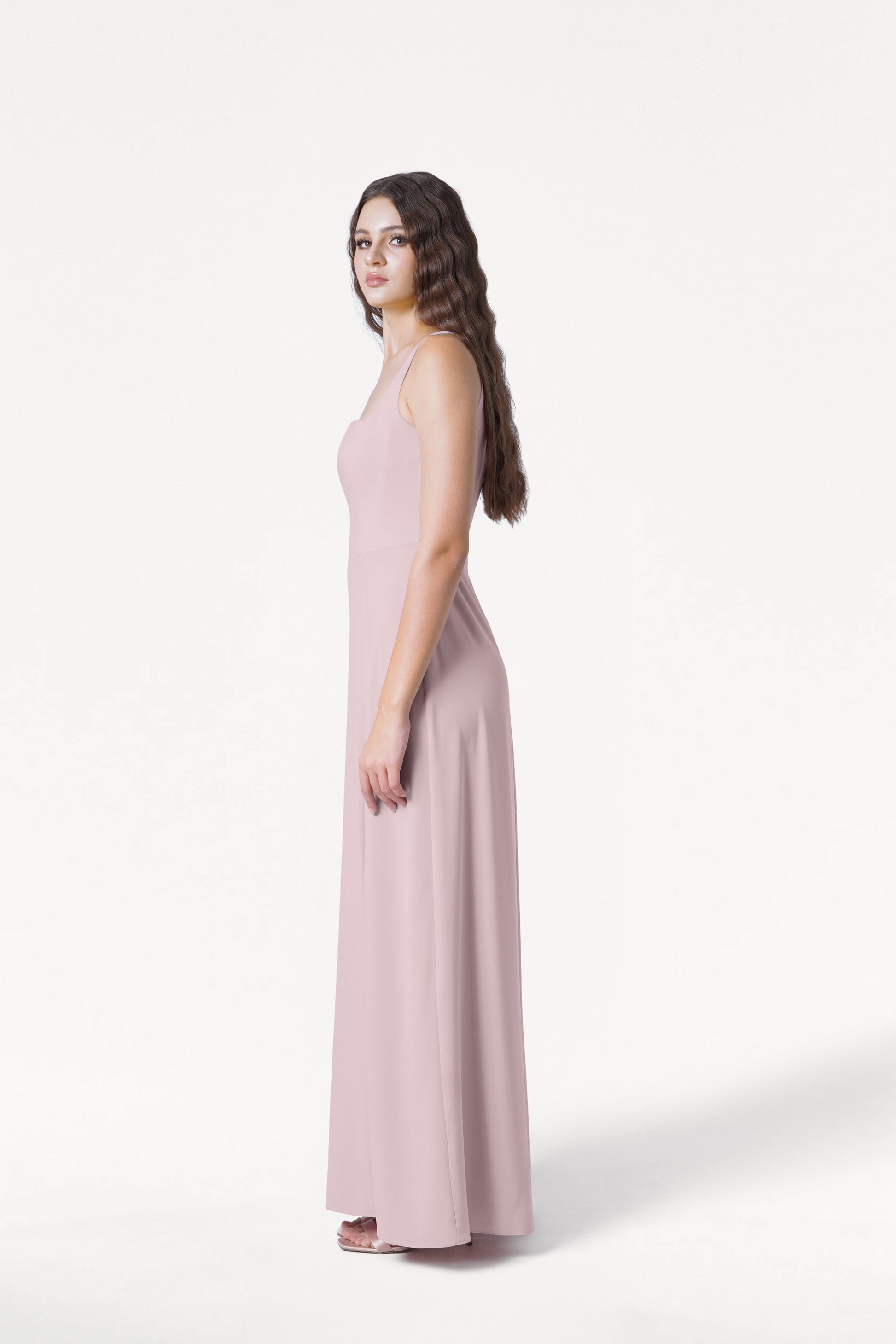 A woman with long wavy brown hair stands in profile wearing the TH&TH Helena Bridesmaid Dress in Smoked Blush and white heels, set against a plain white background.