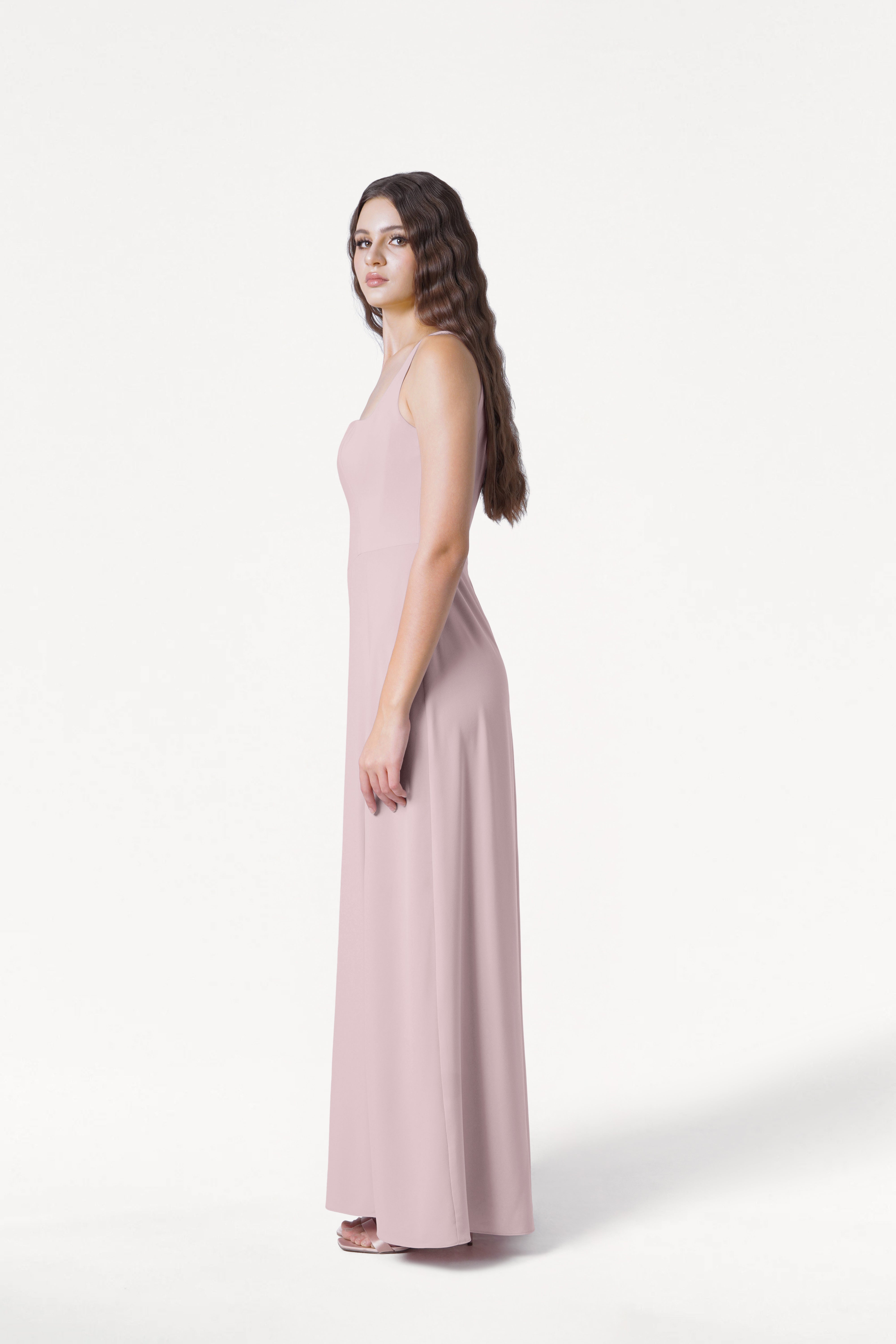 A woman with long wavy brown hair stands in profile wearing the TH&TH Helena Bridesmaid Dress in Smoked Blush and white heels, set against a plain white background.