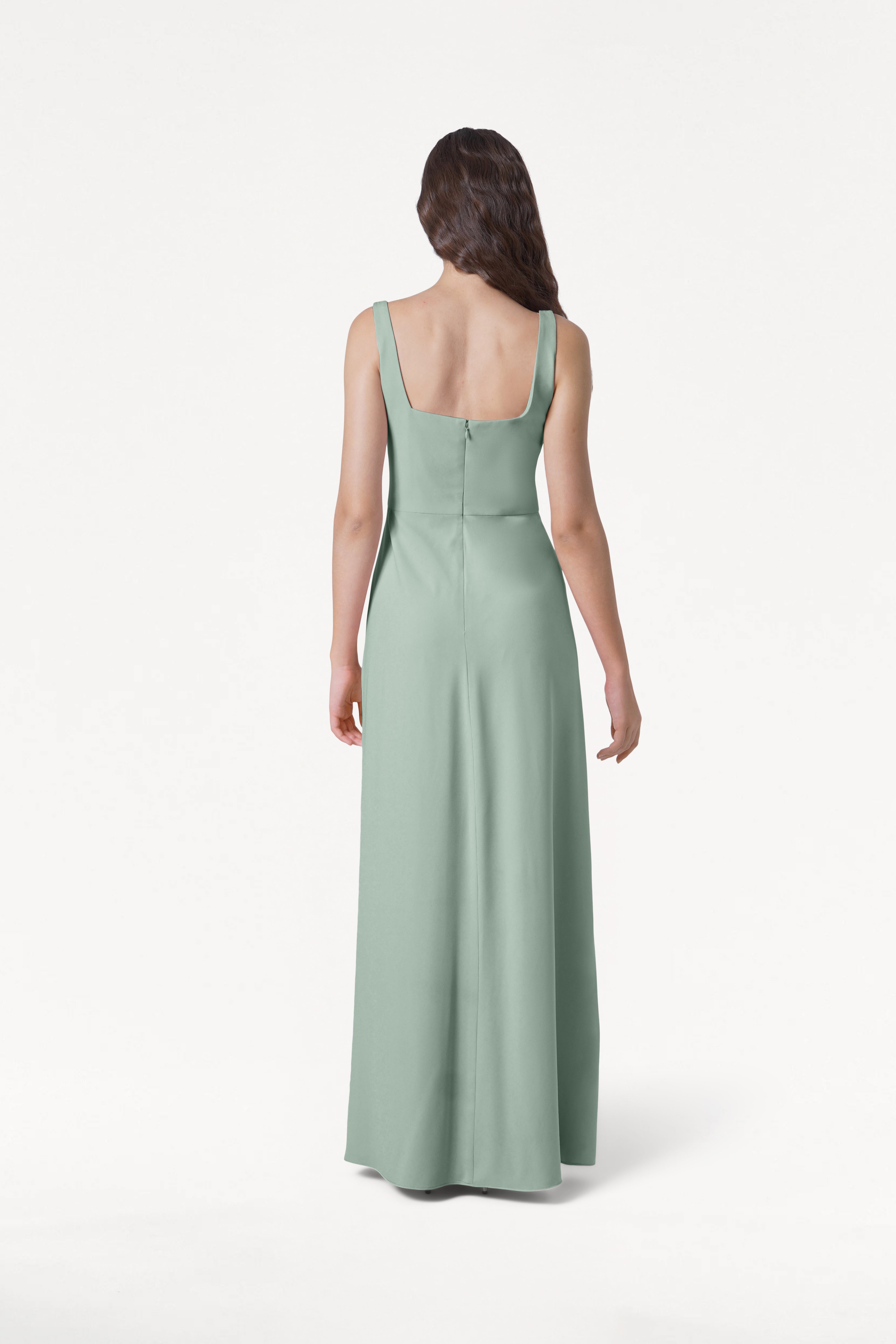 A woman with long, wavy brown hair stands facing away, wearing the TH&TH Helena Bridesmaid Dress in Sage Green, featuring a modern floor-length silhouette, square back, and center seam, set against a plain white background.
