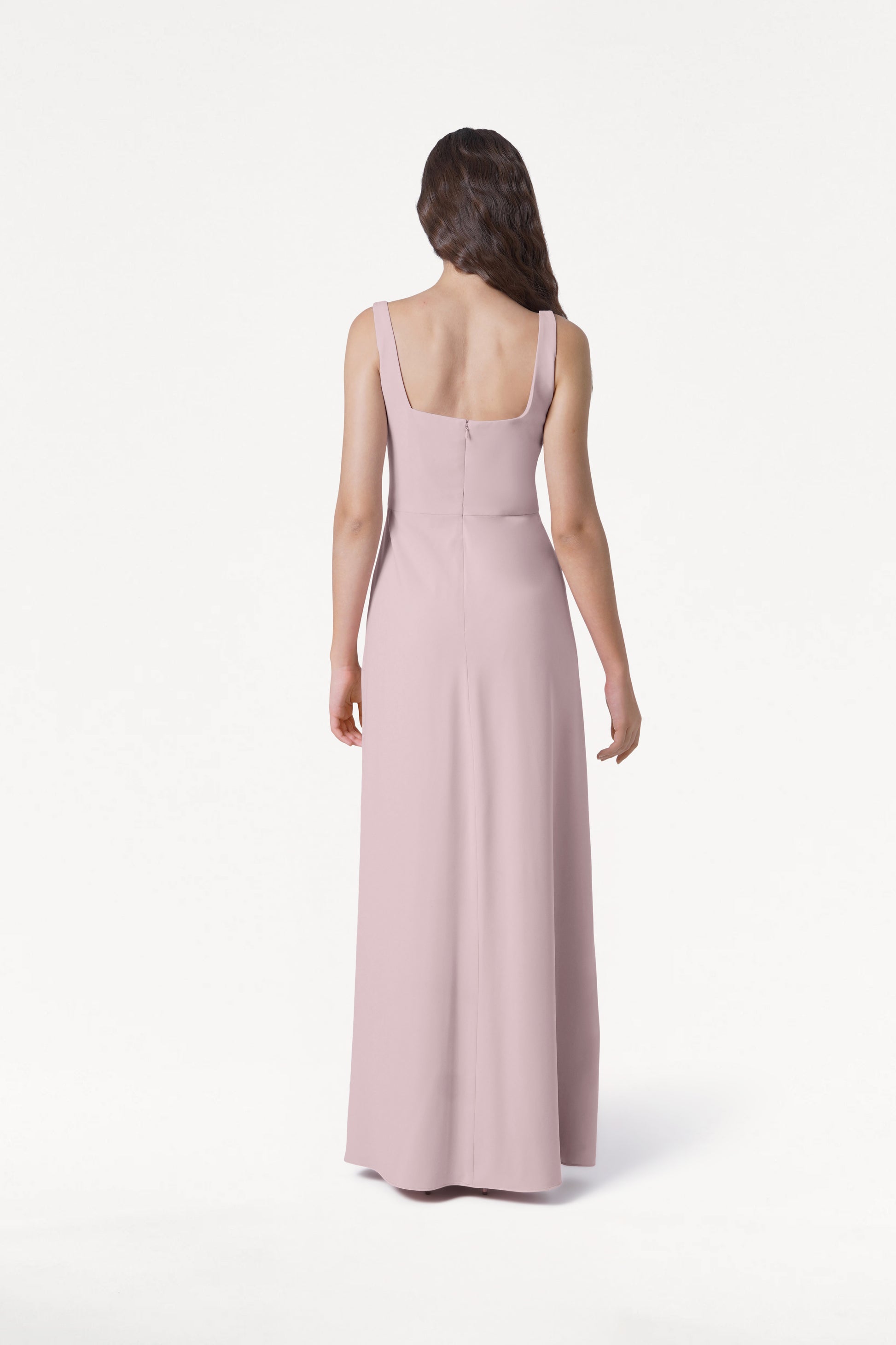 A woman with long, wavy brown hair stands facing away wearing the TH&TH Helena Bridesmaid Dress in Smoked Blush, a sleeveless, floor-length modern gown, against a plain white background.