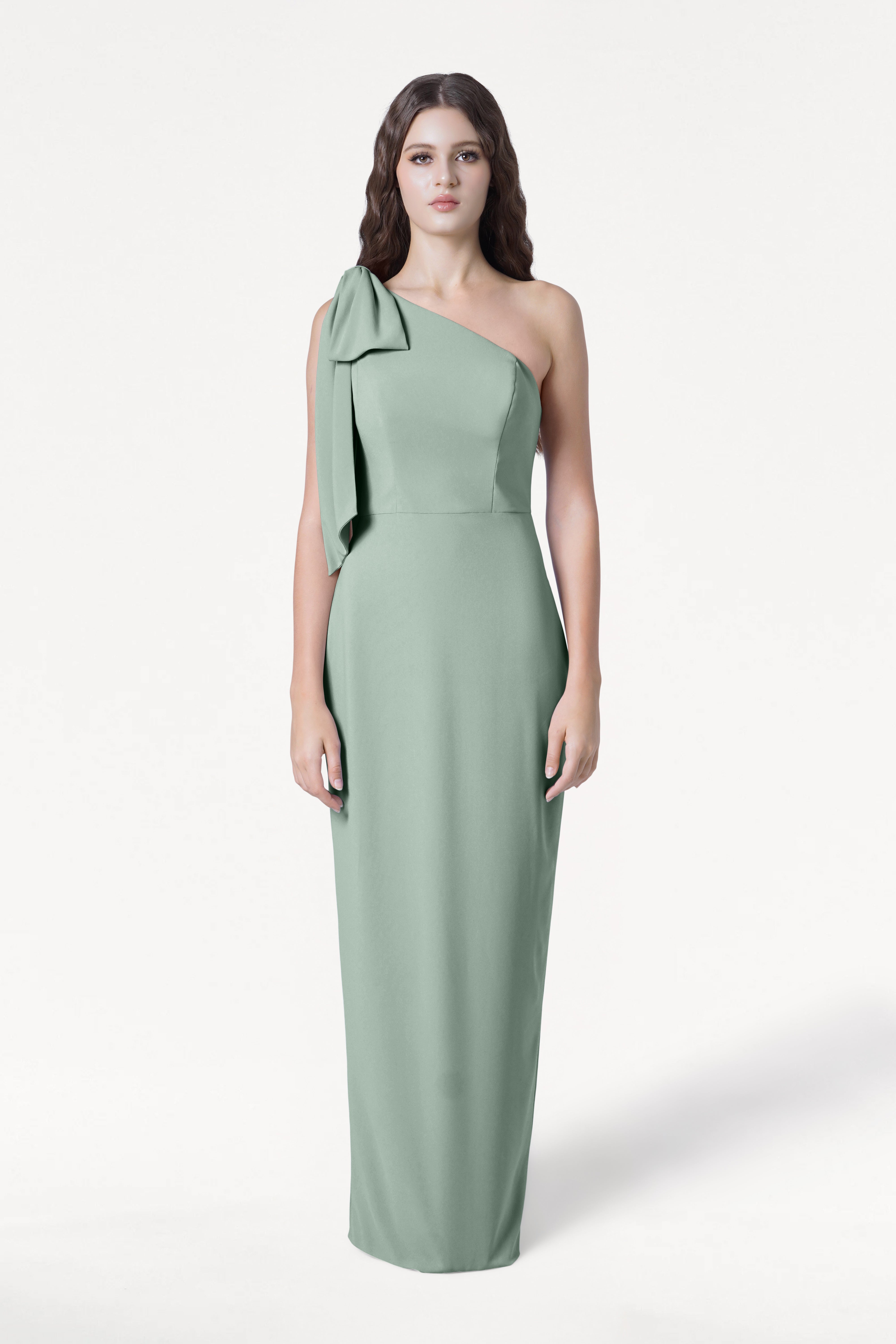 A woman faces forward wearing the TH&TH Valentina Bridesmaid Dress in sage green, featuring size-inclusive fit, a one-shoulder design with bow detail, and modern crepe fabric, set against a plain white background.