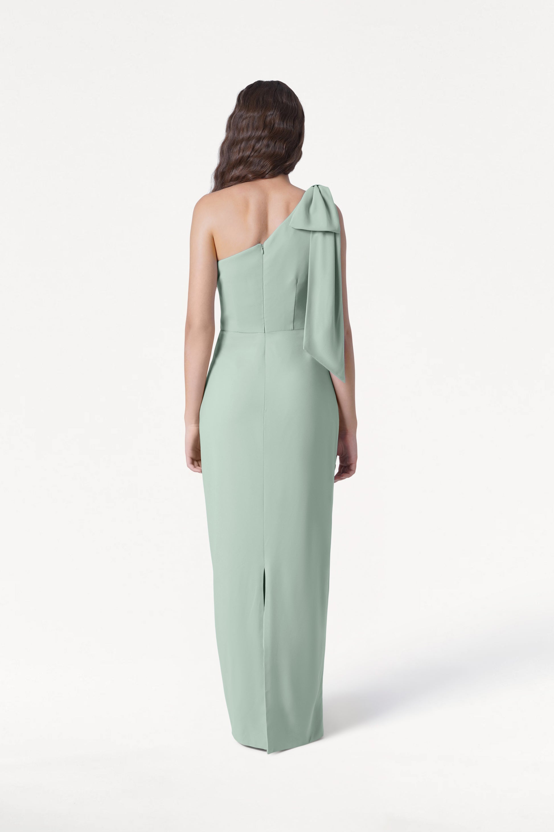 A woman with long wavy hair faces away, modeling the TH&TH Valentina Bridesmaid Dress in Sage Green—a size-inclusive, modern crepe one-shoulder gown with a bow detail—set against a plain white background.