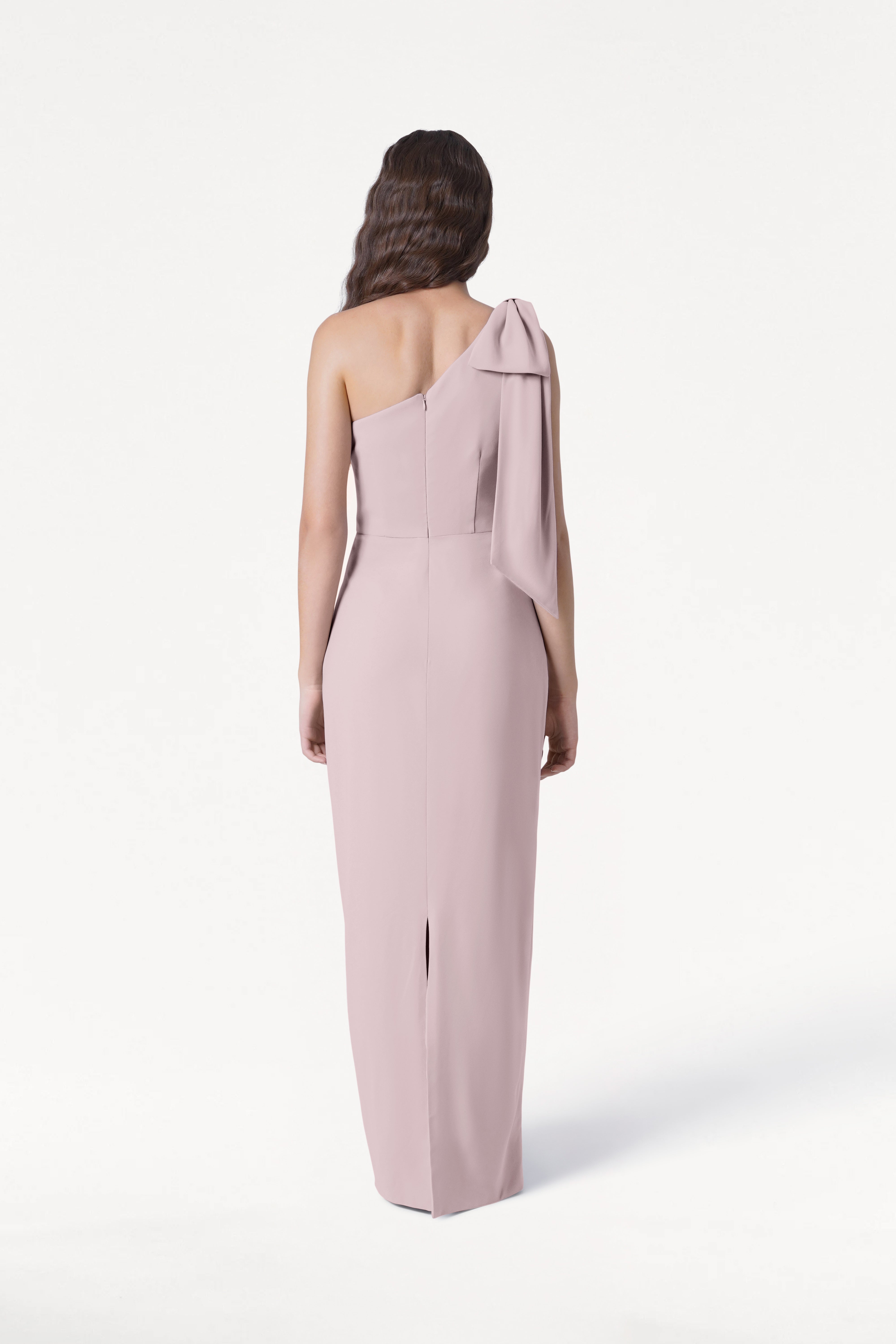 A woman with long, wavy brown hair stands facing away against a white background, wearing the TH&TH Valentina Bridesmaid Dress in Smoked Blush—featuring crepe fabric, a one-shoulder bow, and back slit.