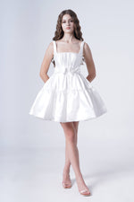 A young woman with long wavy hair models the TH&TH Bloom Bridal Mini Dress in Ivory, a short sleeveless satin twill piece with a flared skirt and floral waist detail, posing hands behind her back against a plain white backdrop.