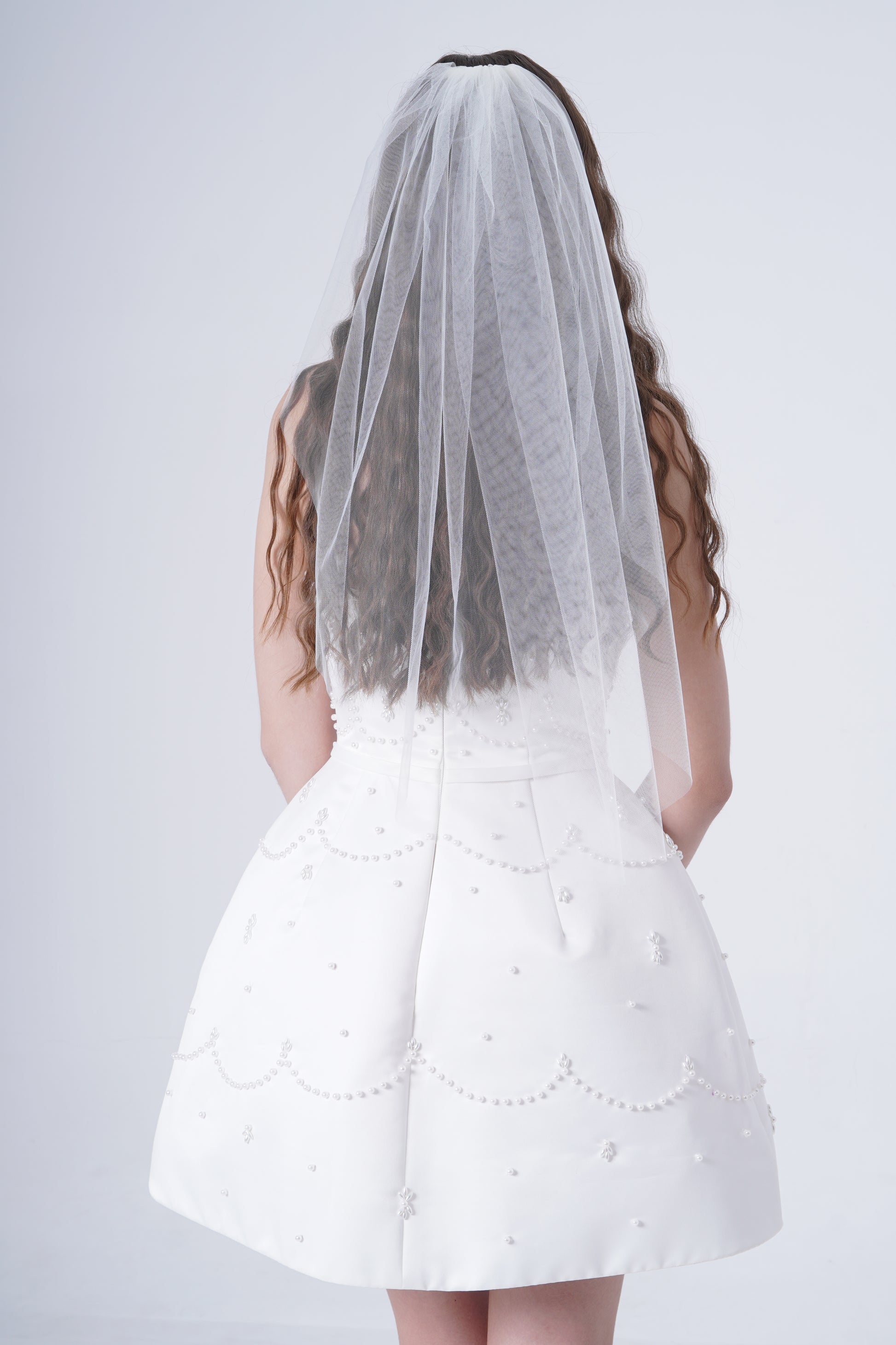 A woman with long wavy hair wears the TH&TH Lucia Plain Veil, styled with a white bridal gown, standing with her back to the camera against a plain light background.