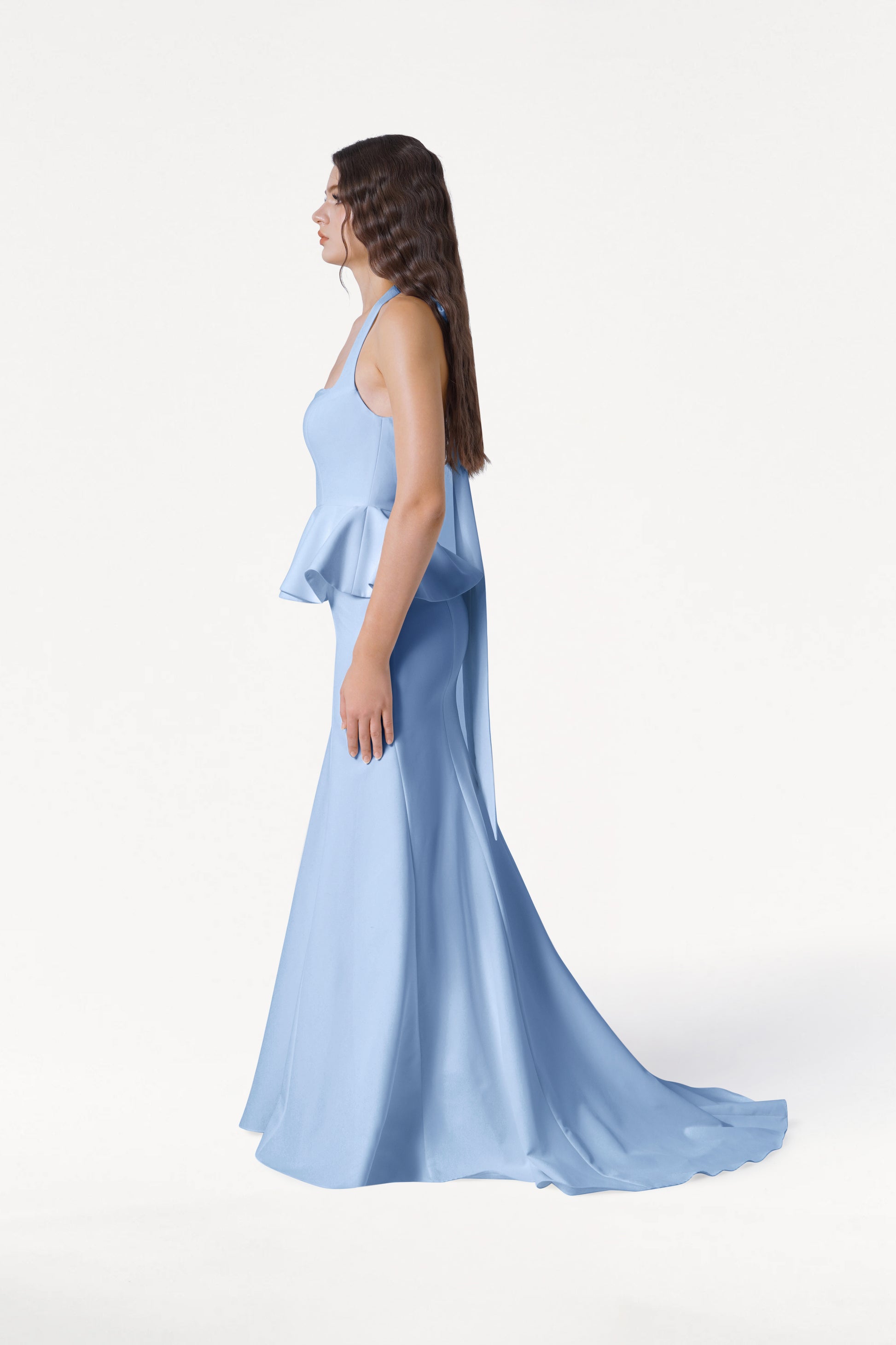 A woman with long brown hair stands in profile against a white background, wearing the TH&TH Peony Bridesmaid Dress in Dusty Blue—a floor-length modern gown with peplum detail and a flowing train.