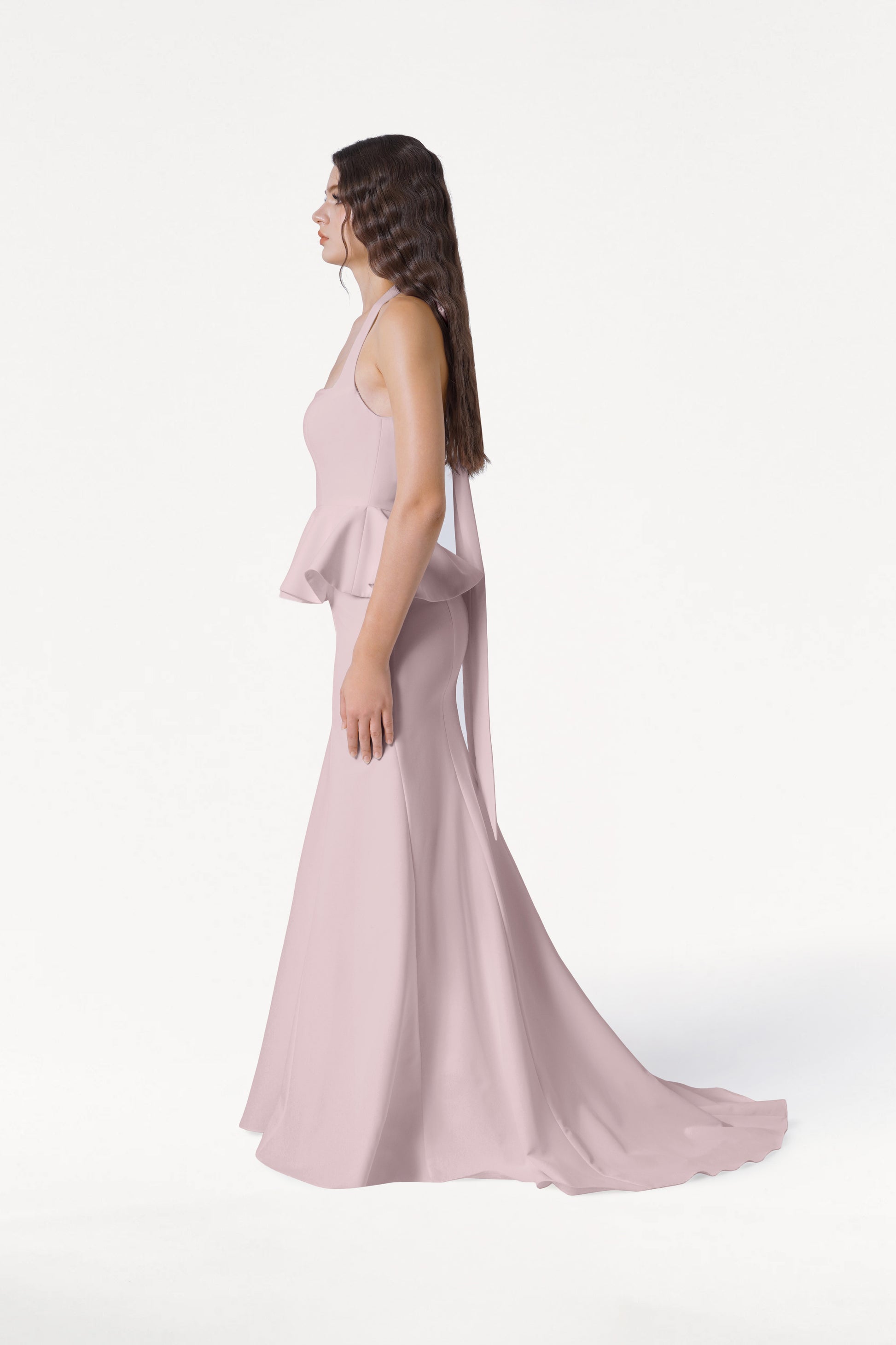 A woman in profile wears the TH&TH Peony Bridesmaid Dress in Smoked Blush, a light pink, sleeveless, floor-length moss crepe gown with peplum detail. Her long, wavy brown hair is worn down against a white background.