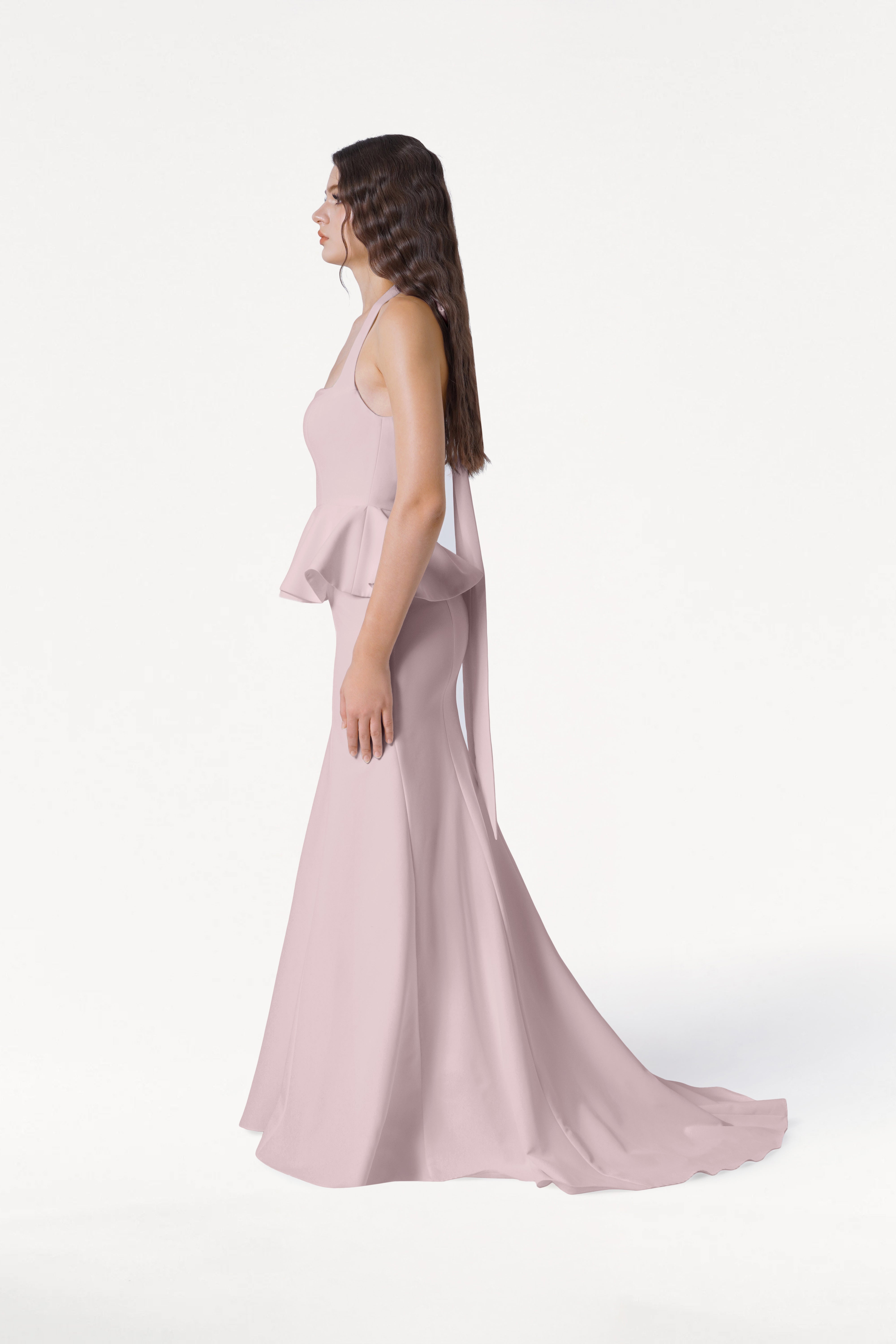 A woman in profile wears the TH&TH Peony Bridesmaid Dress in Smoked Blush, a light pink, sleeveless, floor-length moss crepe gown with peplum detail. Her long, wavy brown hair is worn down against a white background.