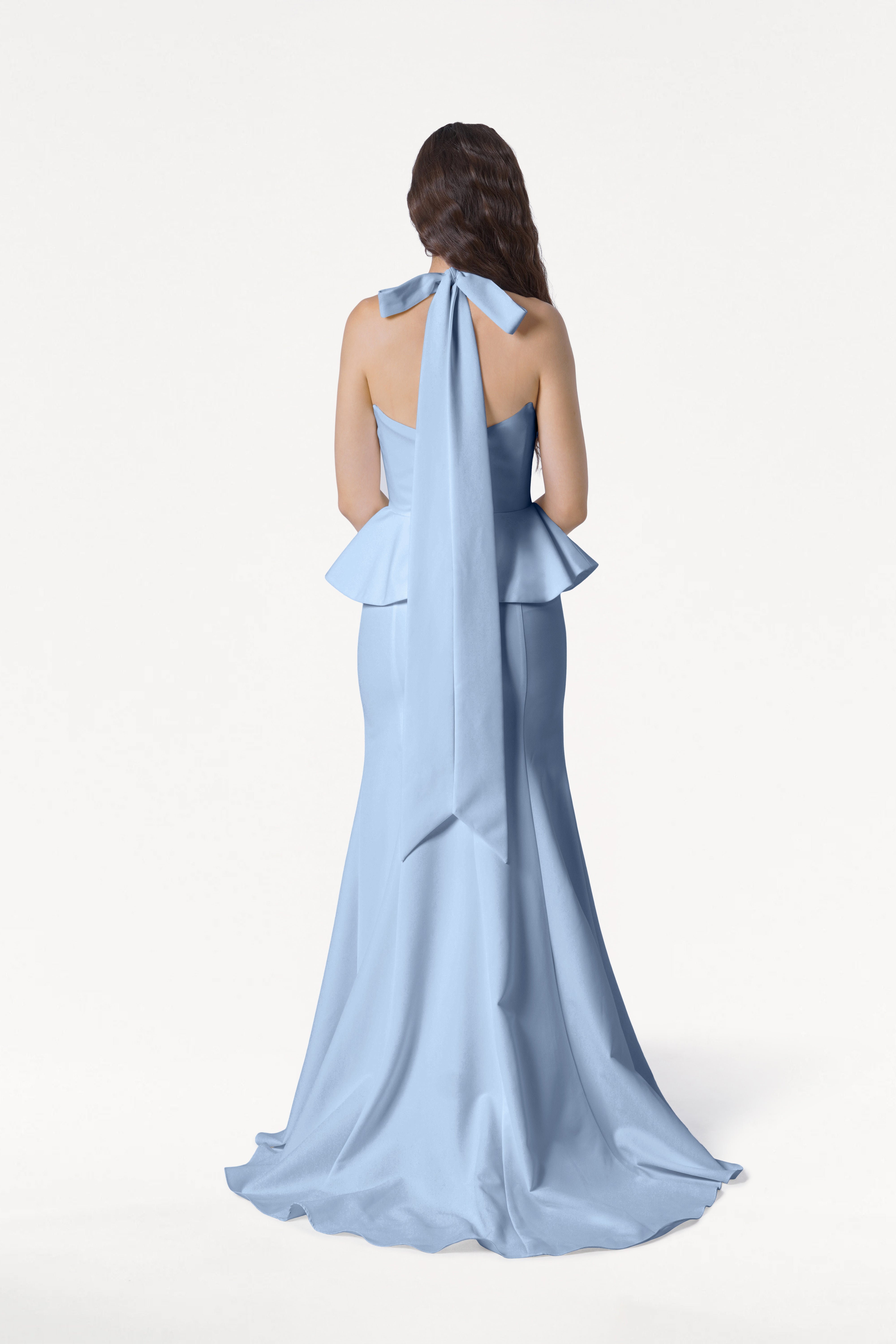A woman with long brown hair faces away, wearing the TH&TH Peony Bridesmaid Dress in Dusty Blue, featuring a floor-length silhouette, moss crepe fabric, peplum waist, and a large neck bow with long tails. Background is plain white.