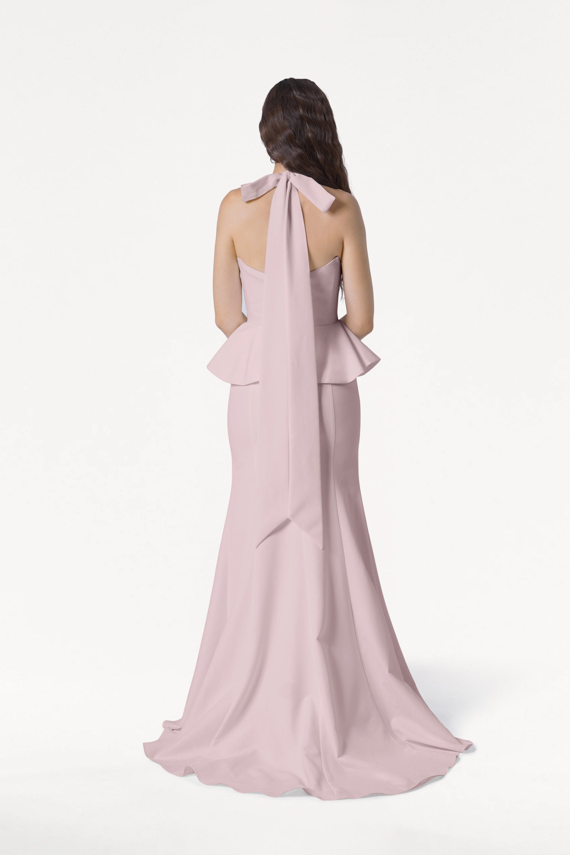 A woman with long brown hair stands facing away, wearing the TH&TH Peony Bridesmaid Dress in Smoked Blush—a modern strapless gown with a ruffled peplum waist and a long bow at the back. The background is plain white.