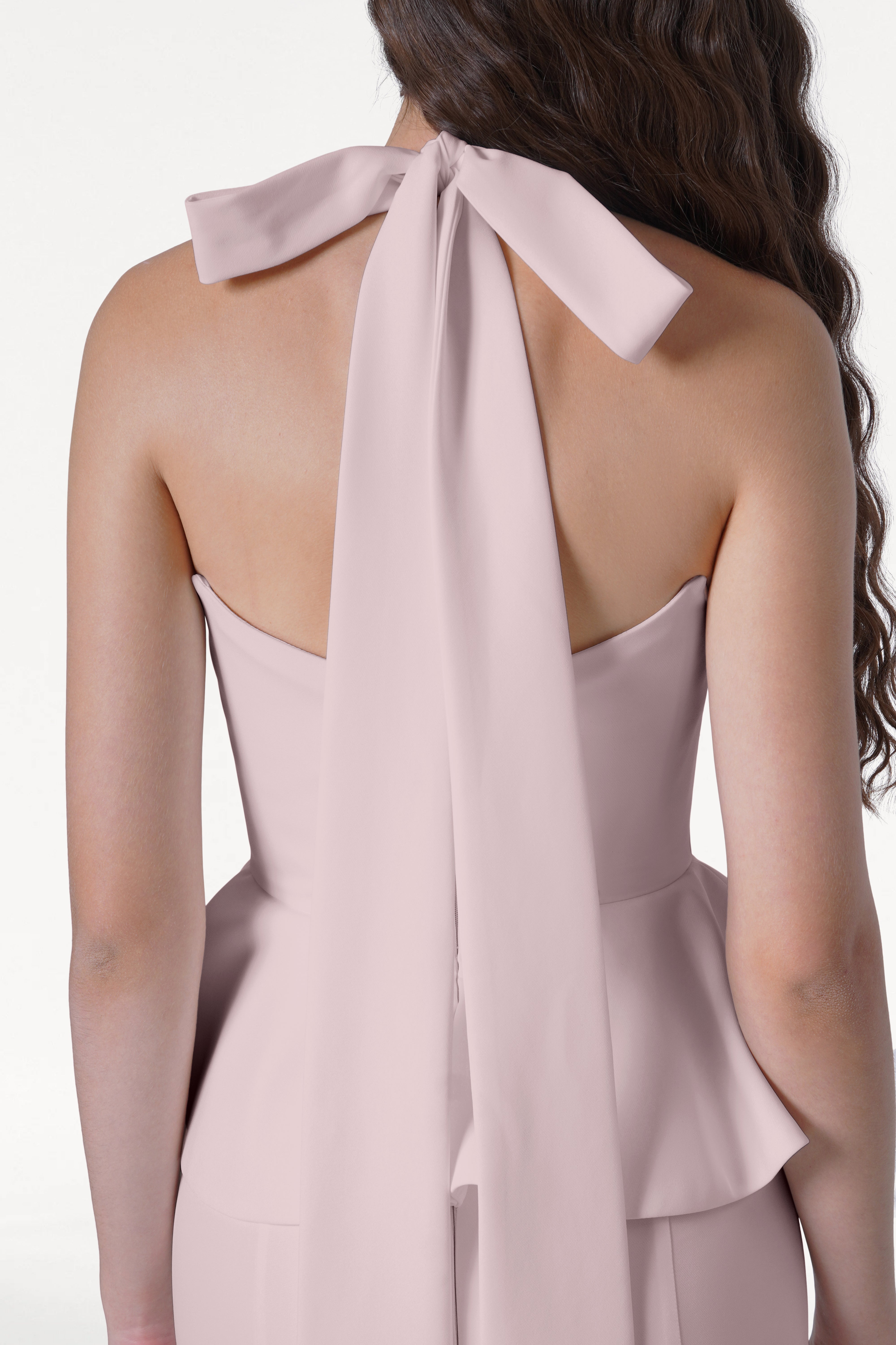 A woman with long, wavy brown hair is shown from the back wearing the TH&TH Peony Bridesmaid Dress in Smoked Blush, a strapless modern gown featuring elegant draping and a large flowing bow at the neck.