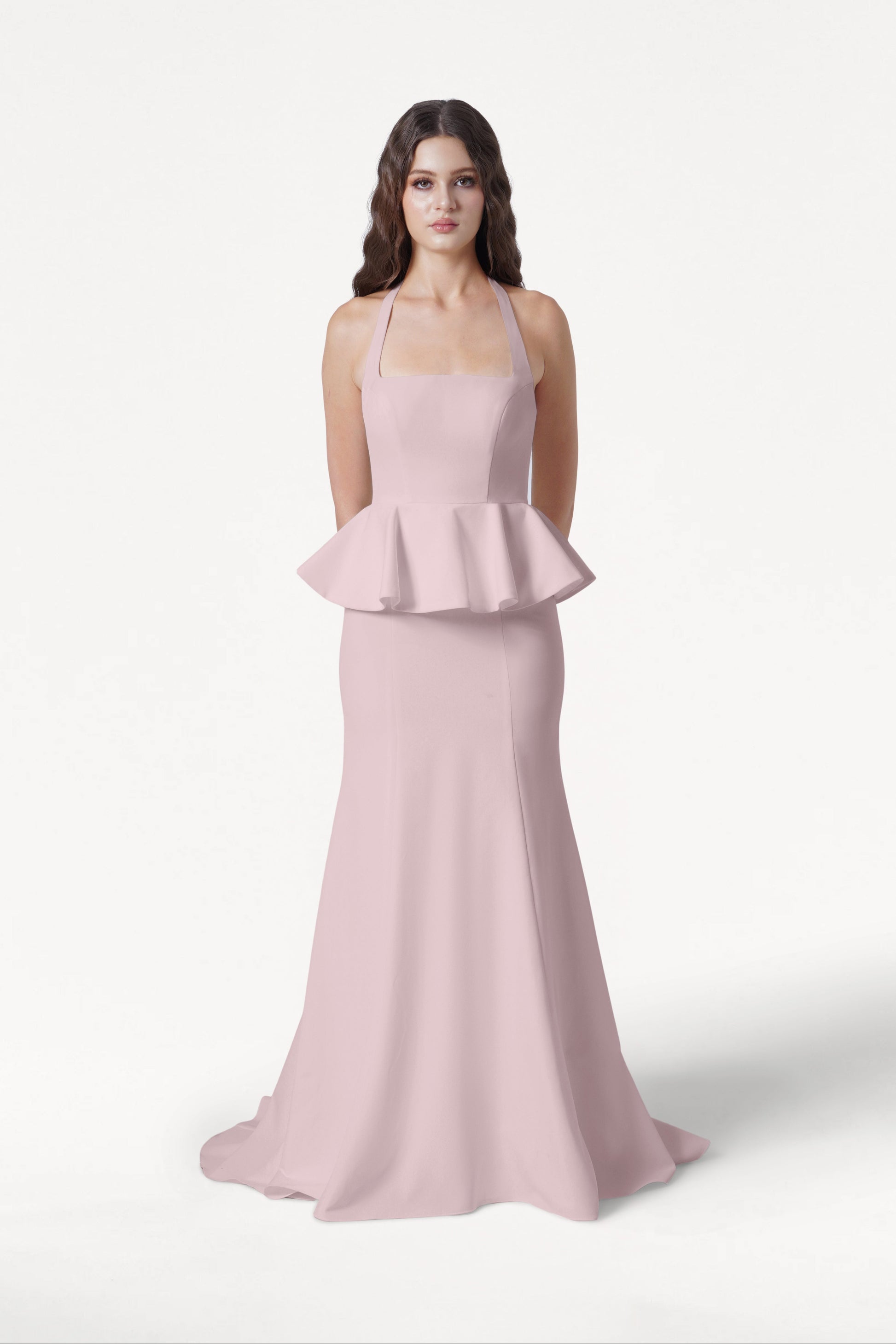 A woman with long brown hair stands against a white background, wearing the TH&TH Peony Bridesmaid Dress in Smoked Blush—a sleeveless gown with a peplum waist and floor-length skirt. Her hands are behind her back.