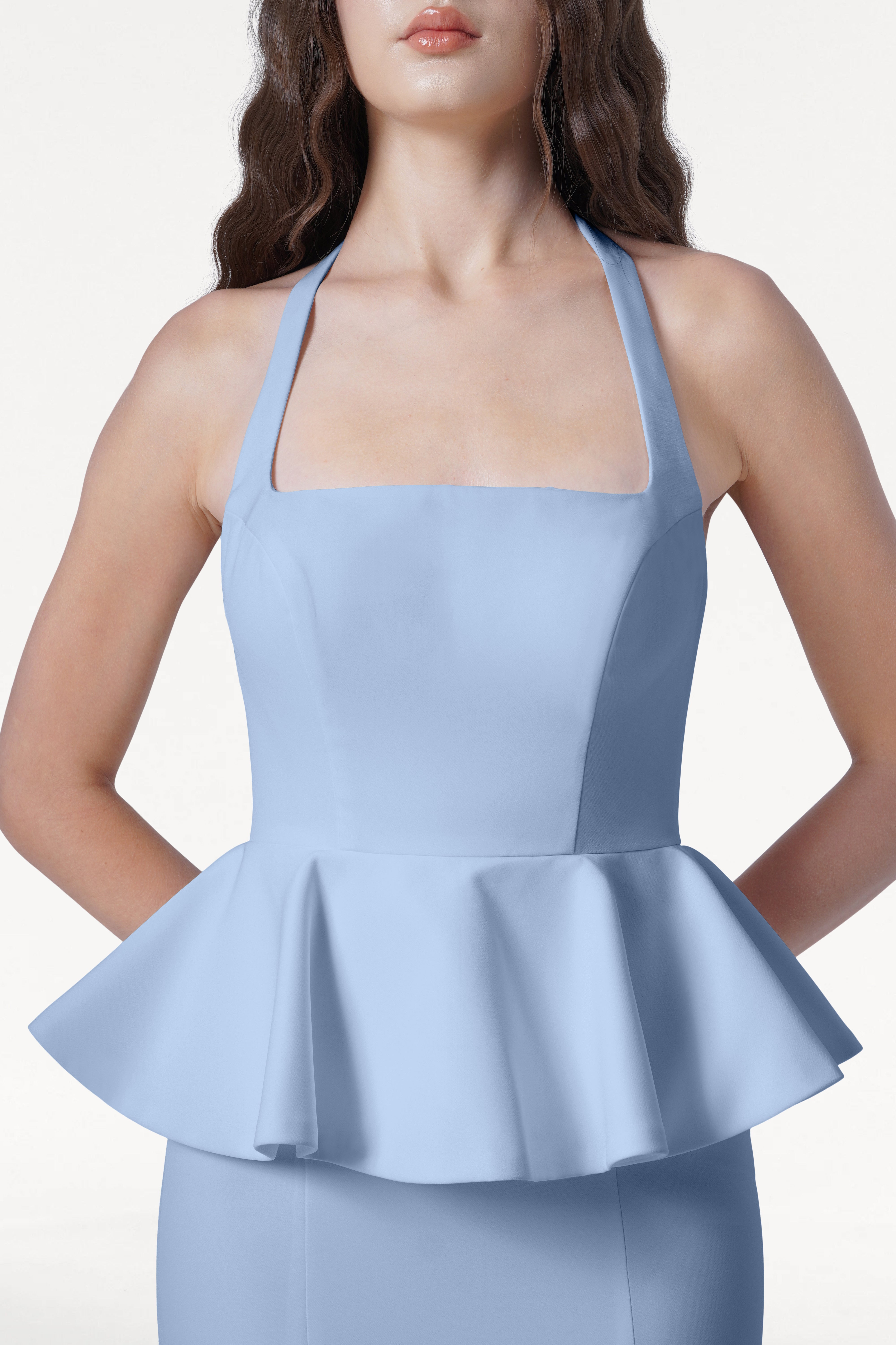 A woman models the Peony Bridesmaid Dress in Dusty Blue by TH&TH, a sleeveless peplum design with a square neckline and halter straps, shown from mid-torso up against a plain white background.