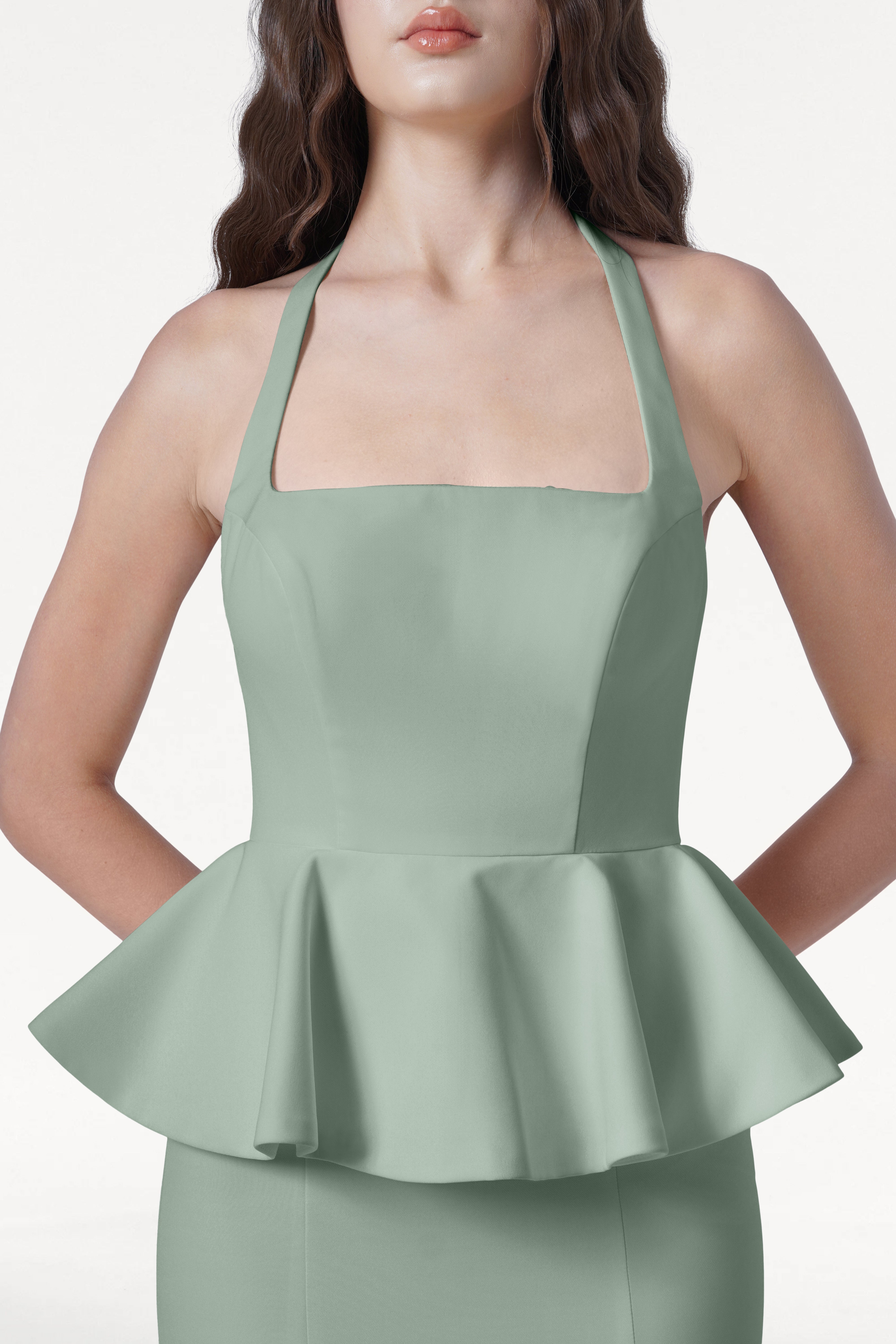 A woman wears the TH&TH Peony Bridesmaid Dress in sage green, a sleeveless gown with a square neckline and halter straps, standing against a plain white background.