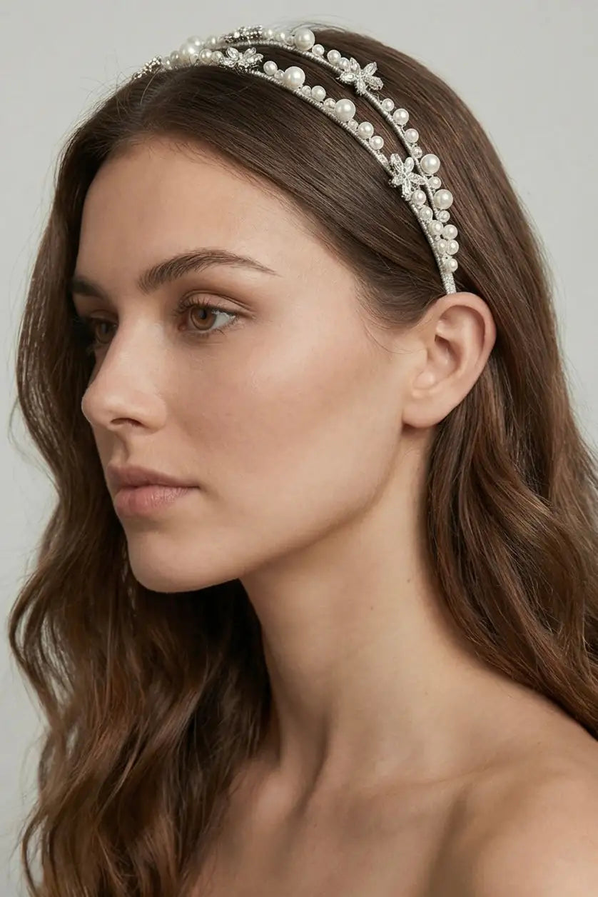 A woman with long, wavy brown hair wears the TH&TH Alta Double Crystal Headband, an elegant bridal accessory. She appears from the shoulders up with a neutral expression against a plain background.