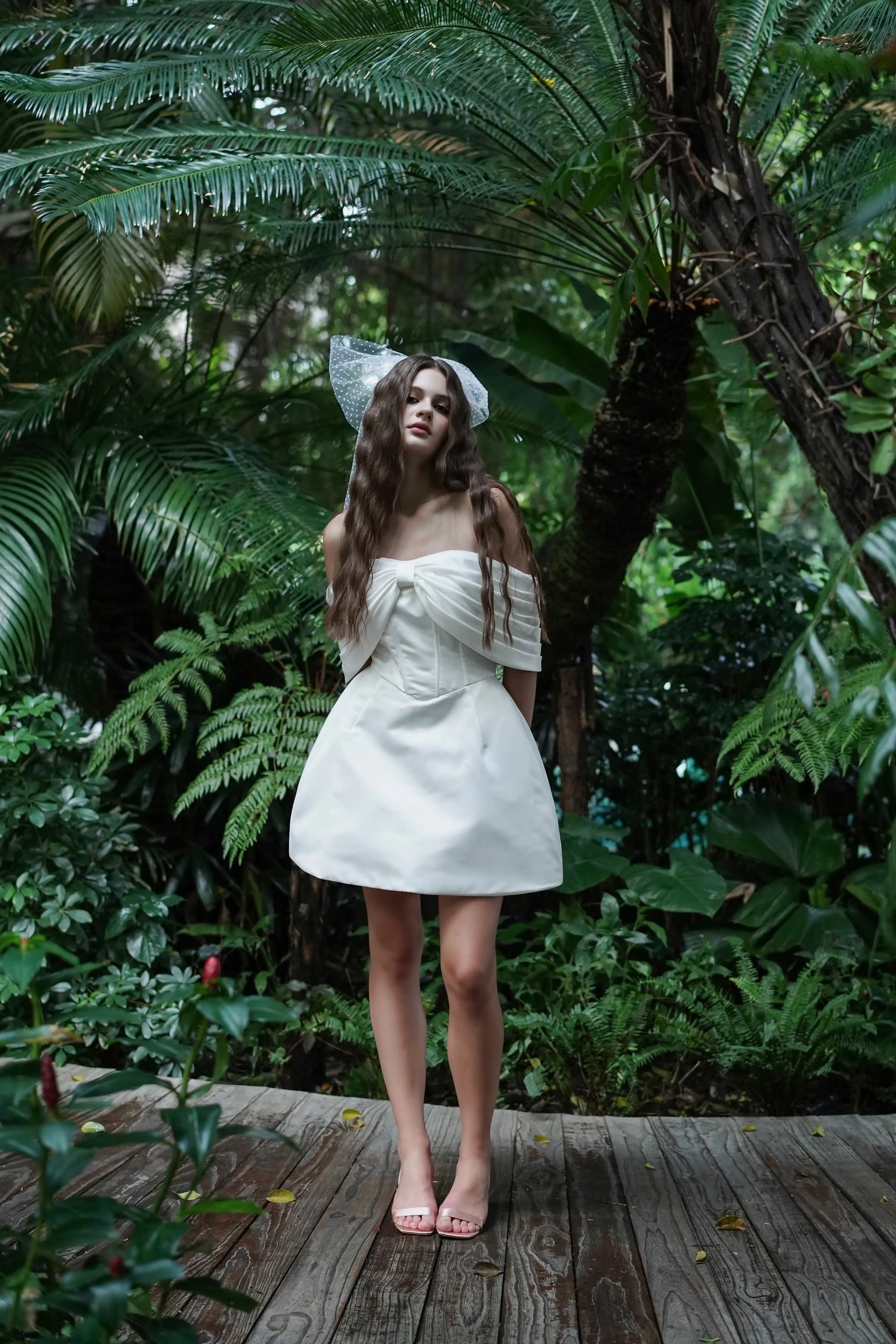 A woman wearing an ivory strapless dress with bow detail and white bow veil in hair standing in the jungle.