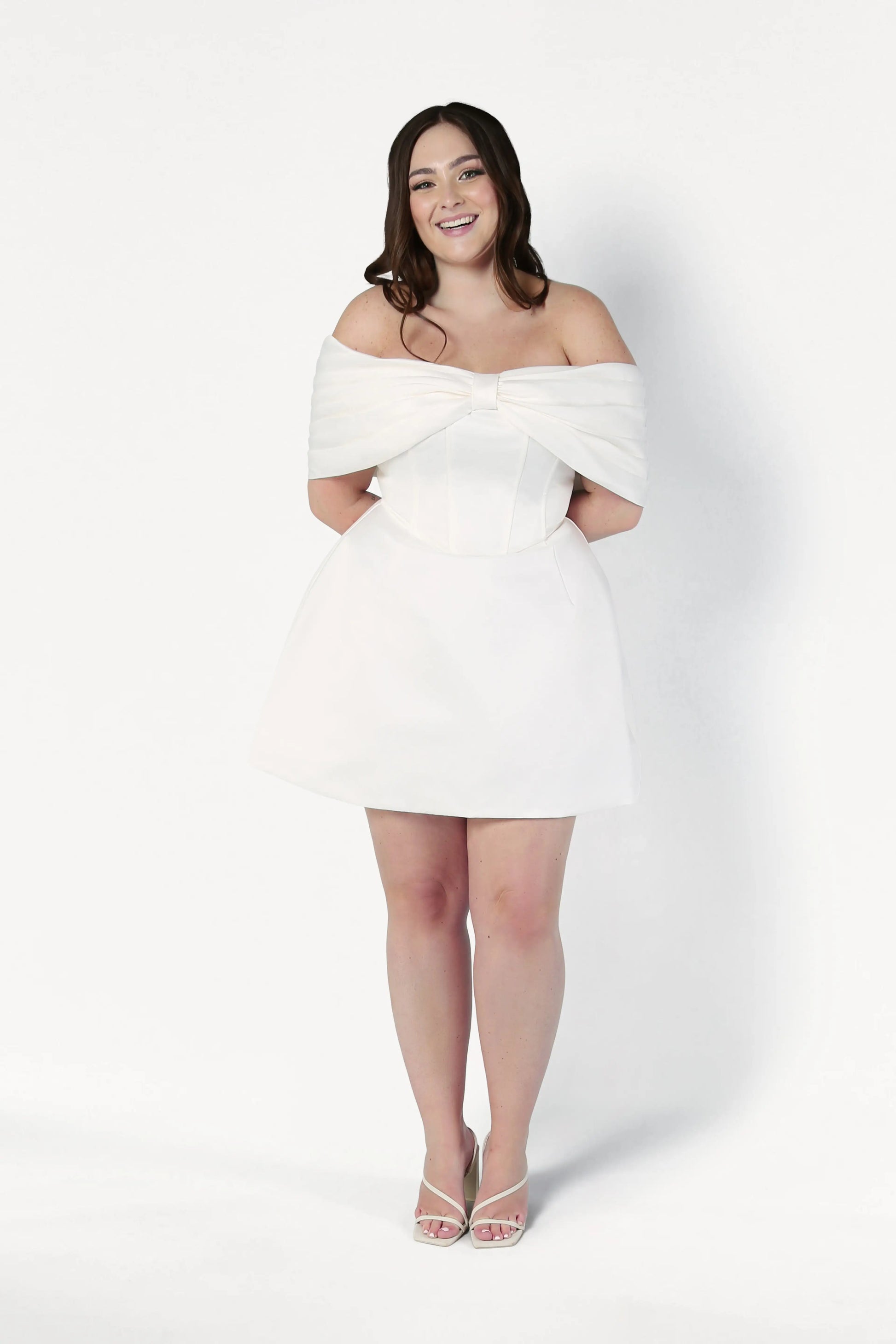 A woman with long brown hair smiles while wearing the Aspen Bridal Mini Dress in Ivory by TH&TH and white heels, standing against a plain white background.