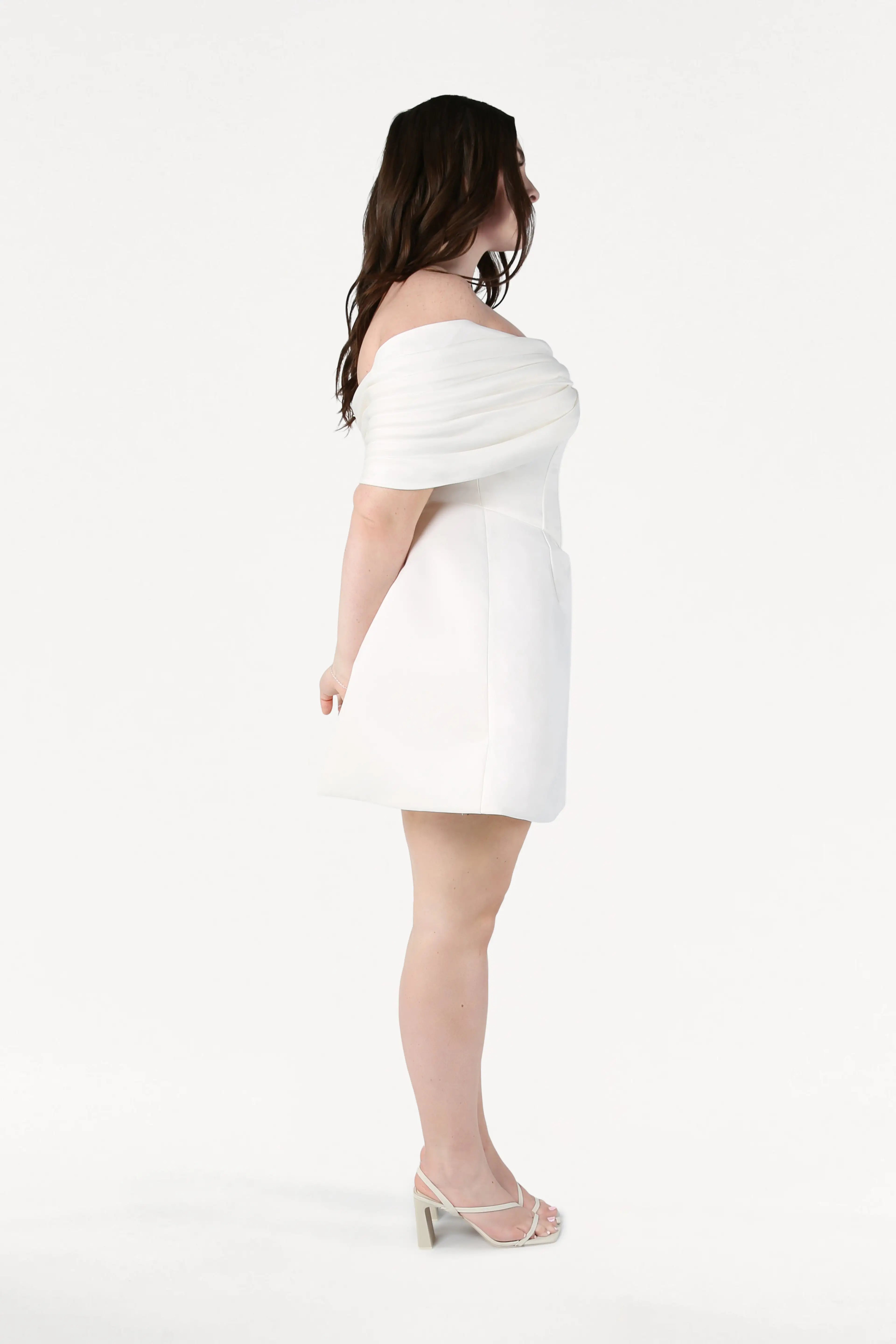 A woman with long brown hair stands in profile against a white background, wearing the TH&TH Aspen Bridal Mini Dress in Ivory with heeled sandals. Her arms are relaxed behind her back.