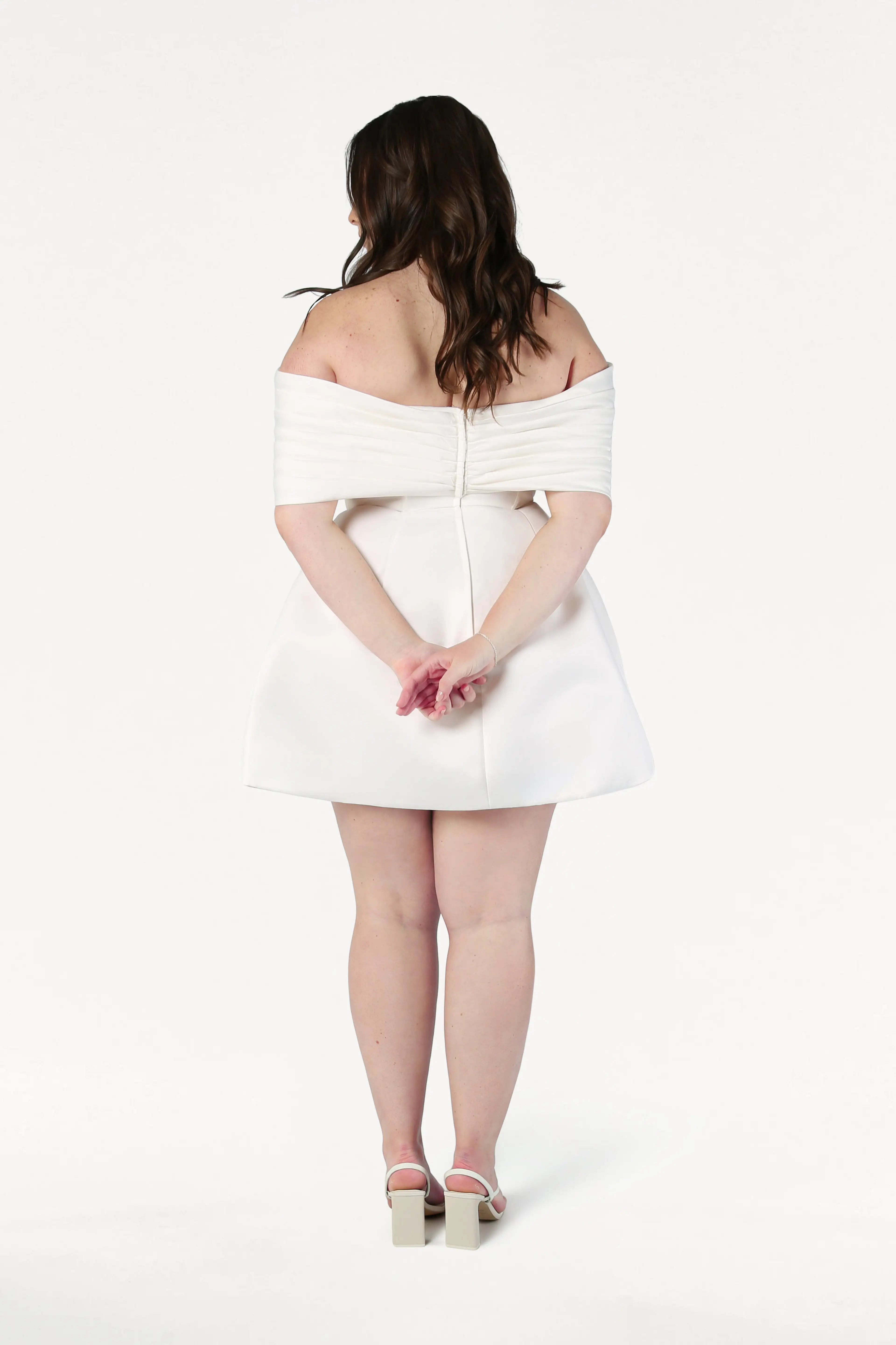 A woman wearing the Aspen Bridal Mini Dress in Ivory by TH&TH stands facing away, arms crossed behind her back, against a plain white background.