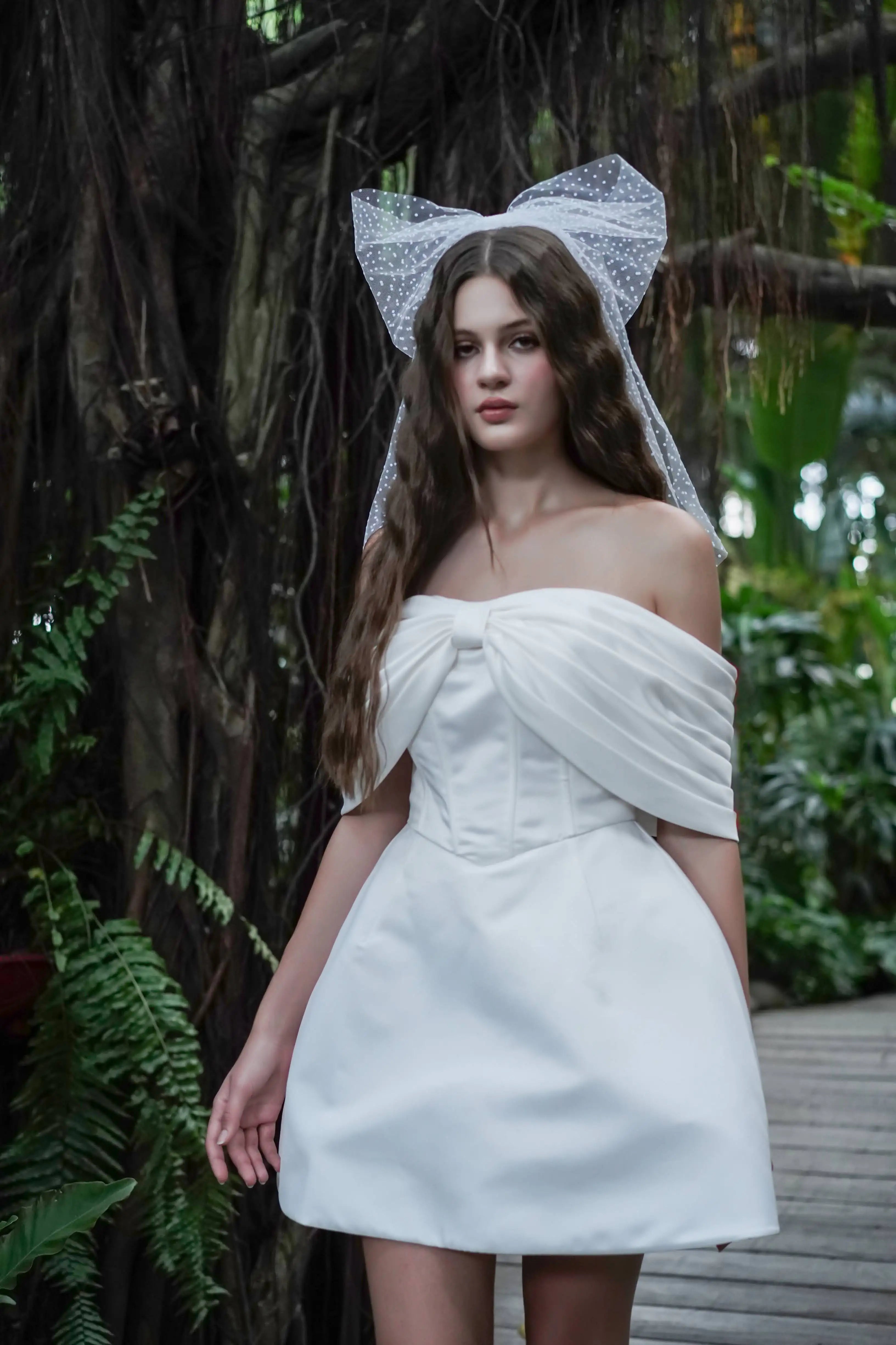 A woman wearing a white strapless dress with bardot sleeves standing in the jungle with bow in her hair.