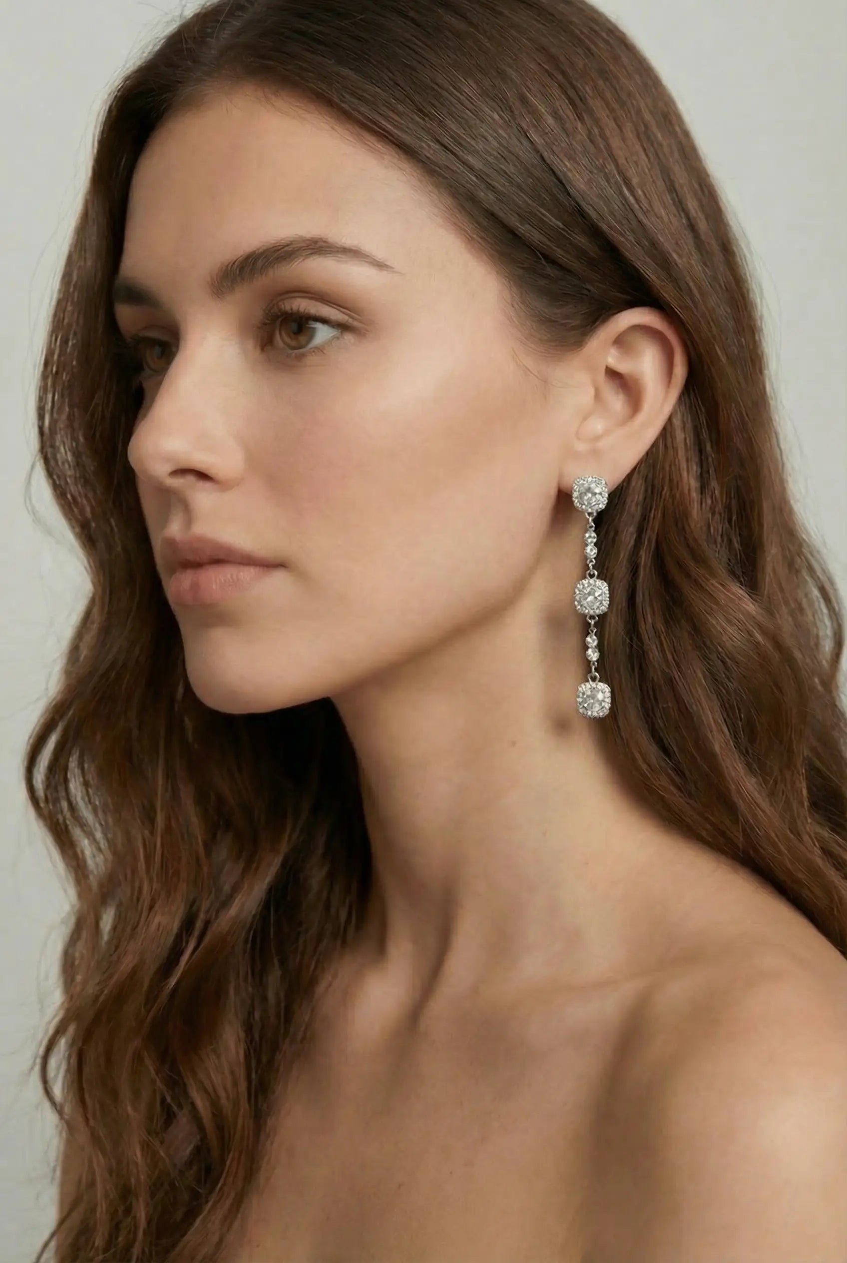 A woman with long brown hair wears TH&TH's Astoria Square Crystal Drop Earrings, featuring three cushion-cut crystal clusters linked by round stones—an exquisite bridal choice—against a light grey background.