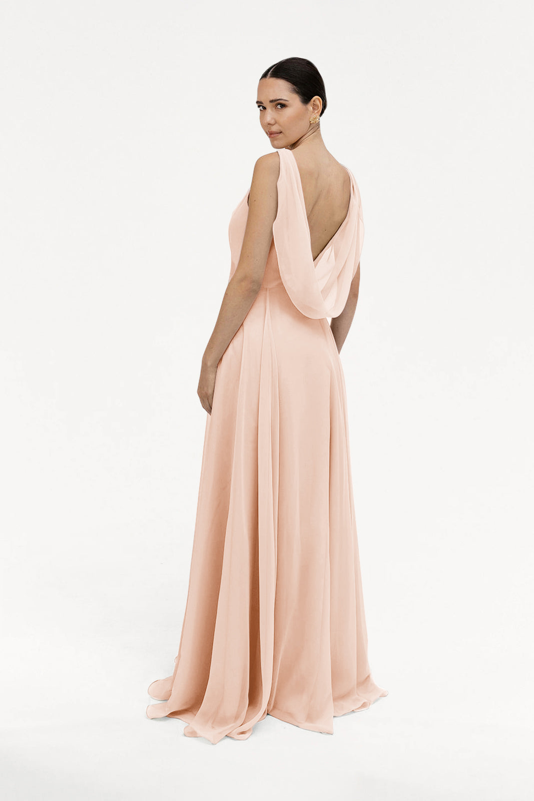 A woman, her dark hair sleekly pulled back, stands against a white background in the TH&TH Athena Bridesmaid Dress in blush pink chiffon, featuring a long, flowing silhouette and a draped cowl back as she looks over her shoulder.