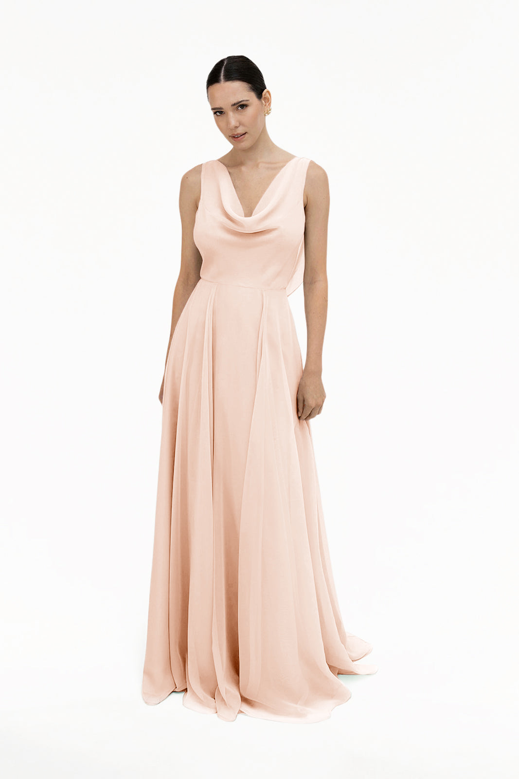 A woman models the TH&TH Athena Bridesmaid Dress in blush pink chiffon, featuring a long, sleeveless design and draped cowl neck, as she stands against a plain white background and looks slightly toward the camera.