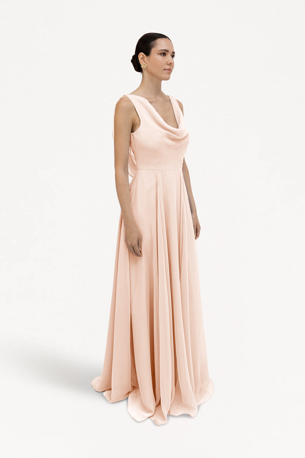 A woman with slicked-back hair stands against a white background, wearing the TH&TH Athena Bridesmaid Dress in blush pink chiffon—a sleeveless, floor-length gown featuring a draped cowl neck.