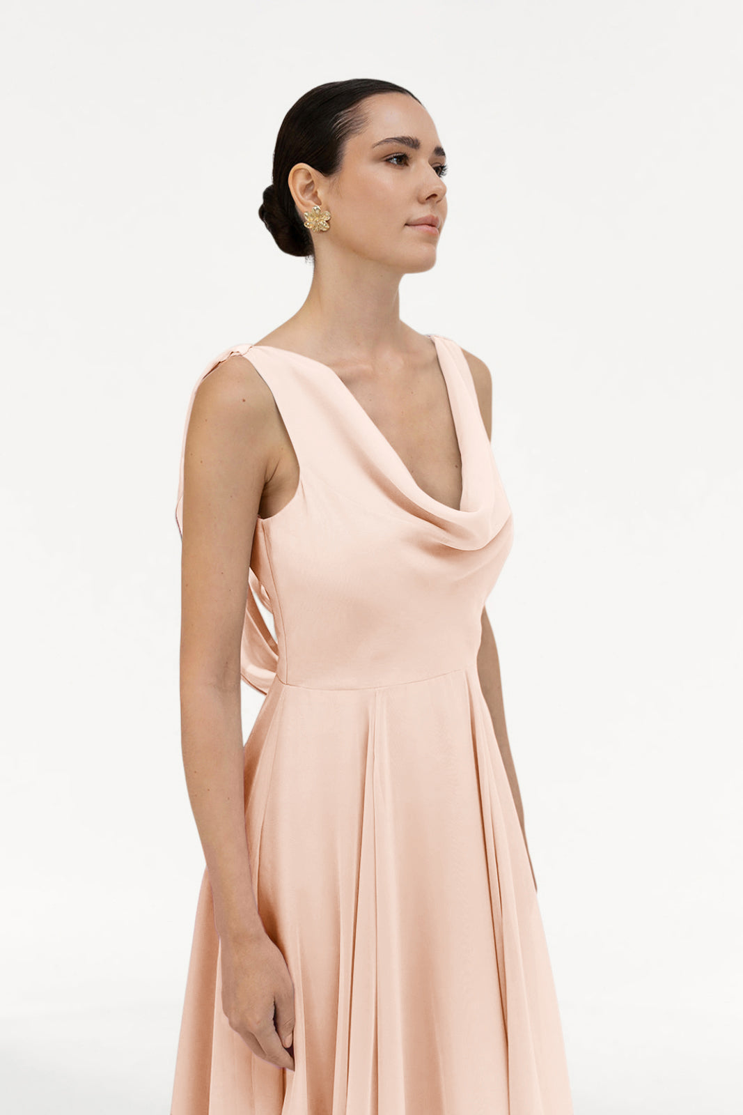 A woman stands against a white background in the TH&TH Athena Bridesmaid Dress in blush pink chiffon, featuring a sleeveless design and cowl neckline. Her hair is styled back, and she accessorizes with small gold earrings.