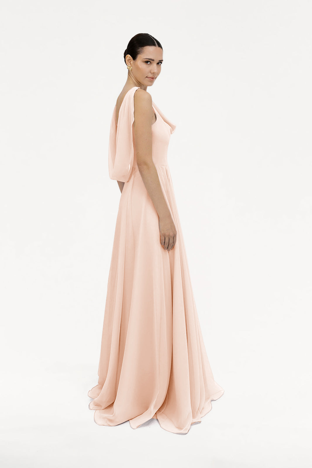 A woman wears the TH&TH Athena Bridesmaid Dress in blush pink chiffon, a floor-length gown with draped sleeves and a cowl neck, posing against a plain white background.