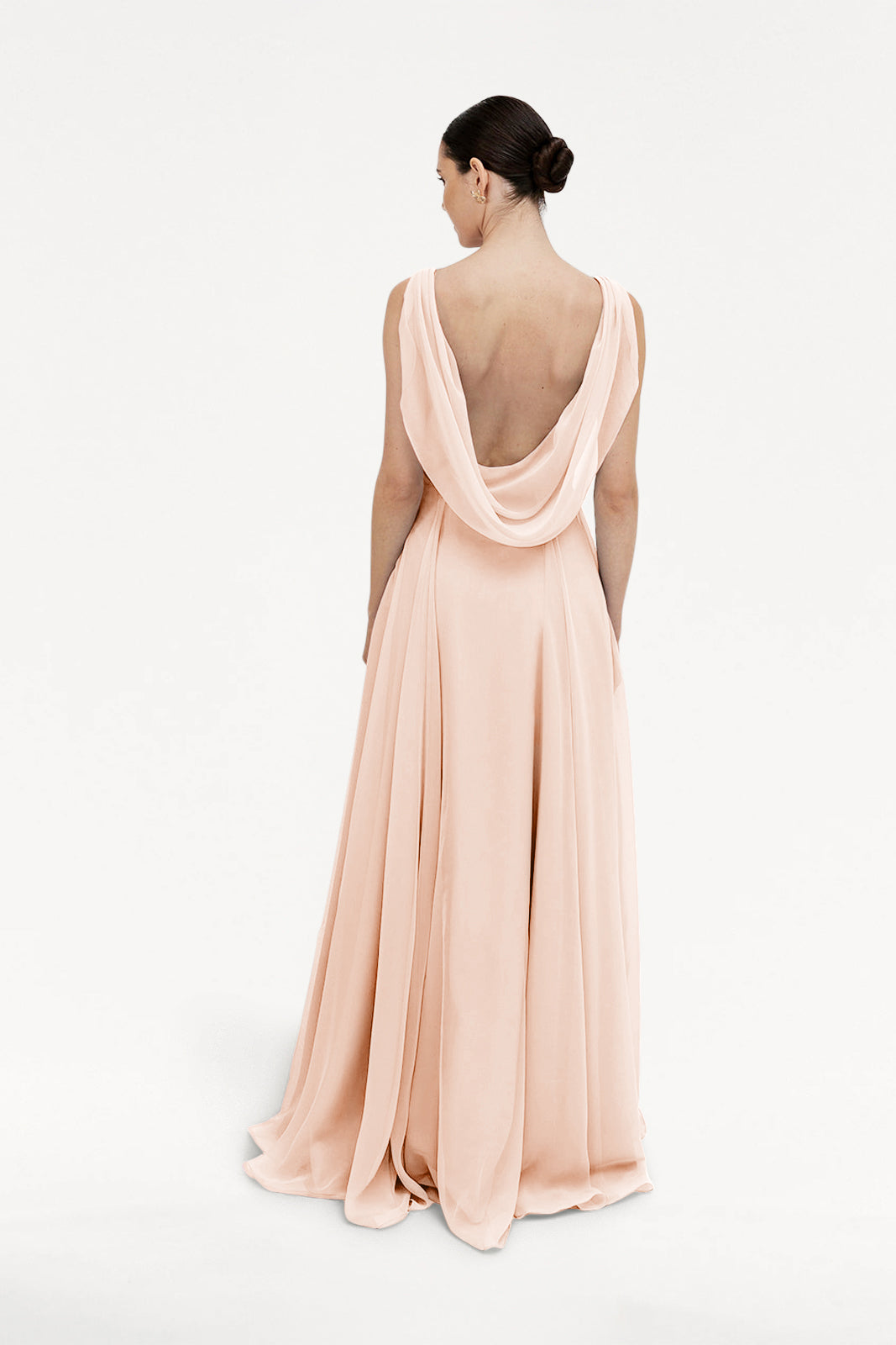 A woman with dark hair in a sleek bun wears the TH&TH Athena Bridesmaid Dress in blush pink chiffon, featuring a cowl neck and draped open back, standing against a plain white background with her back to the camera.