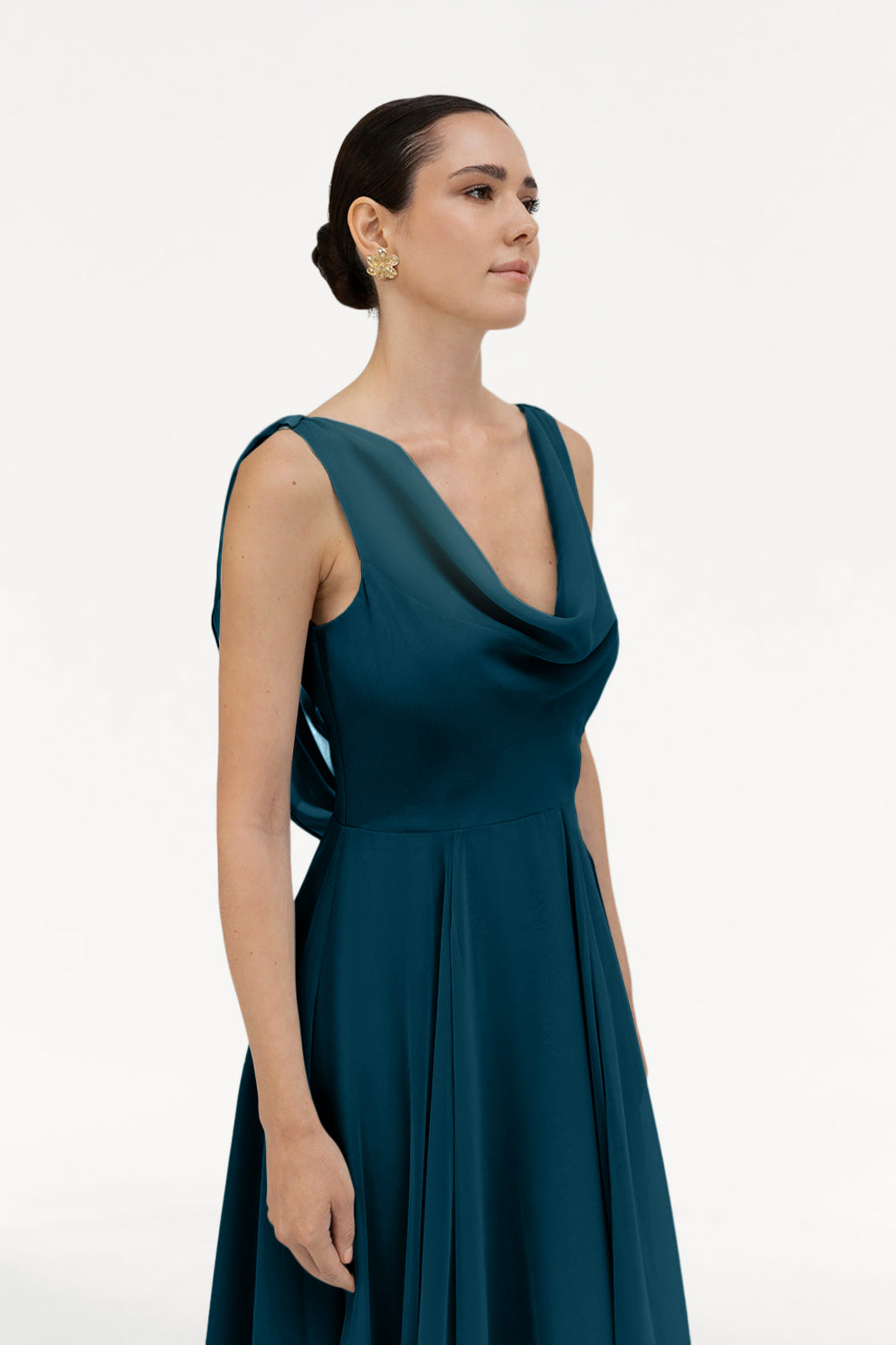 A woman with dark hair in a low bun wears the TH&TH Athena Bridesmaid Dress in Emerald Dawn and gold earrings, radiating timeless elegance against a plain white background.