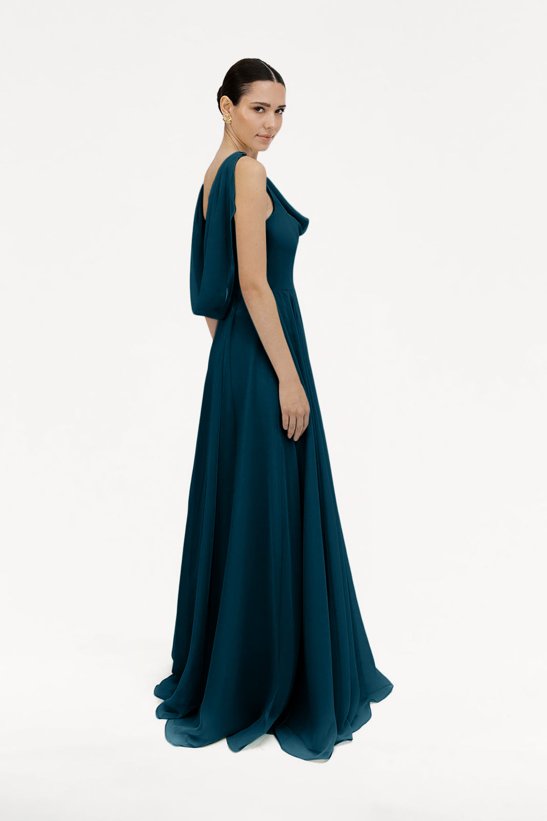 A woman models the TH&TH Athena Bridesmaid Dress in Emerald Dawn, featuring a flowing chiffon silhouette with draped fabric and a cowl neck, standing against a plain white background and radiating timeless elegance.