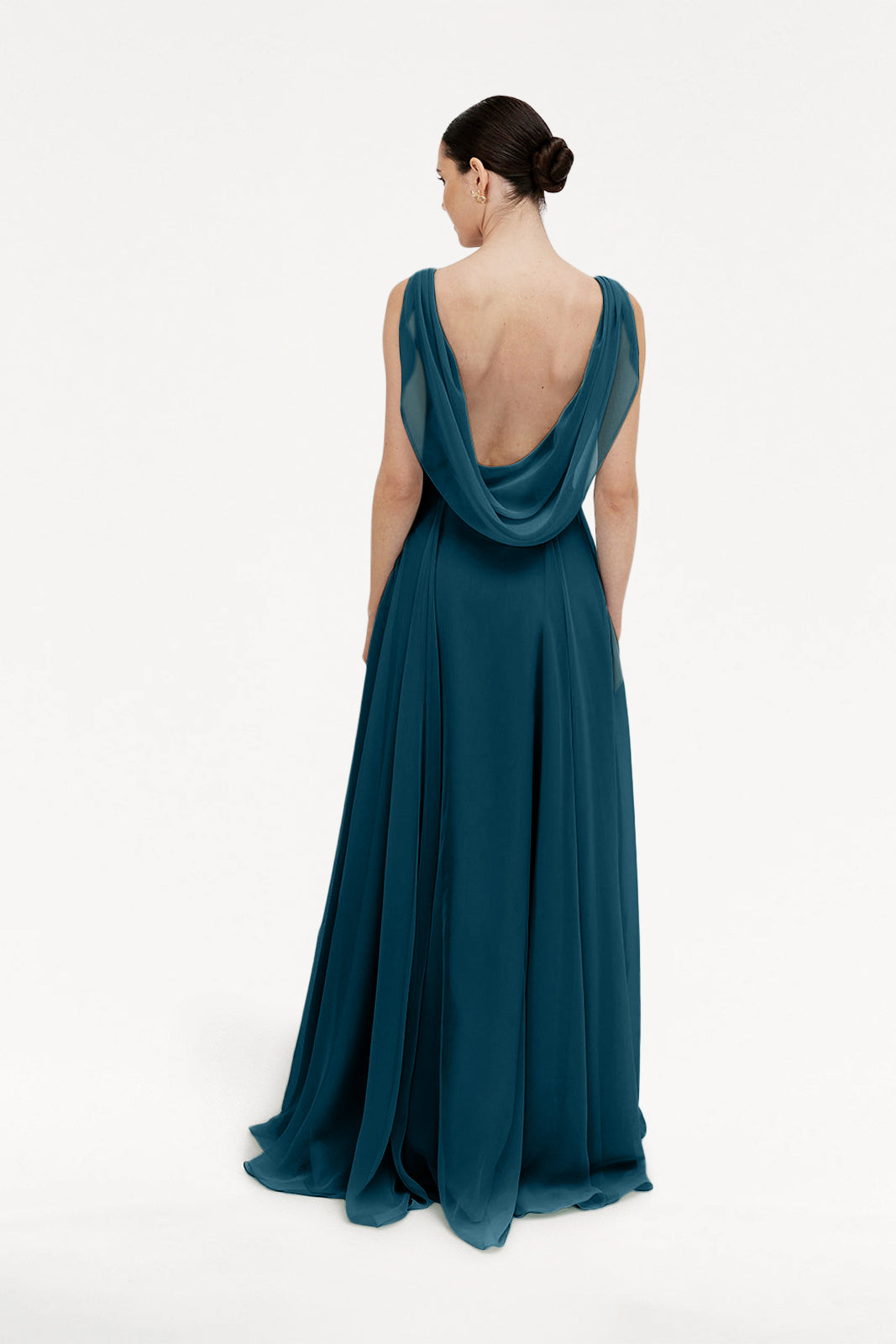 A woman stands with her back to the camera, wearing the TH&TH Athena Bridesmaid Dress in Emerald Dawn—a floor-length chiffon gown with a dramatic low cowl back—her dark hair in a low bun, exuding timeless elegance against a white background.