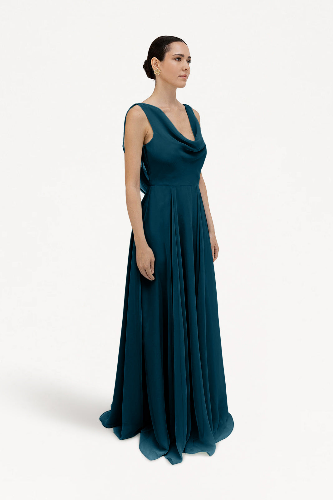 A woman wears the TH&TH Athena Bridesmaid Dress in Emerald Dawn, a sleeveless, floor-length chiffon gown with a draped neckline. Her hair is pulled back as she stands against a plain white background, gazing slightly to the side.